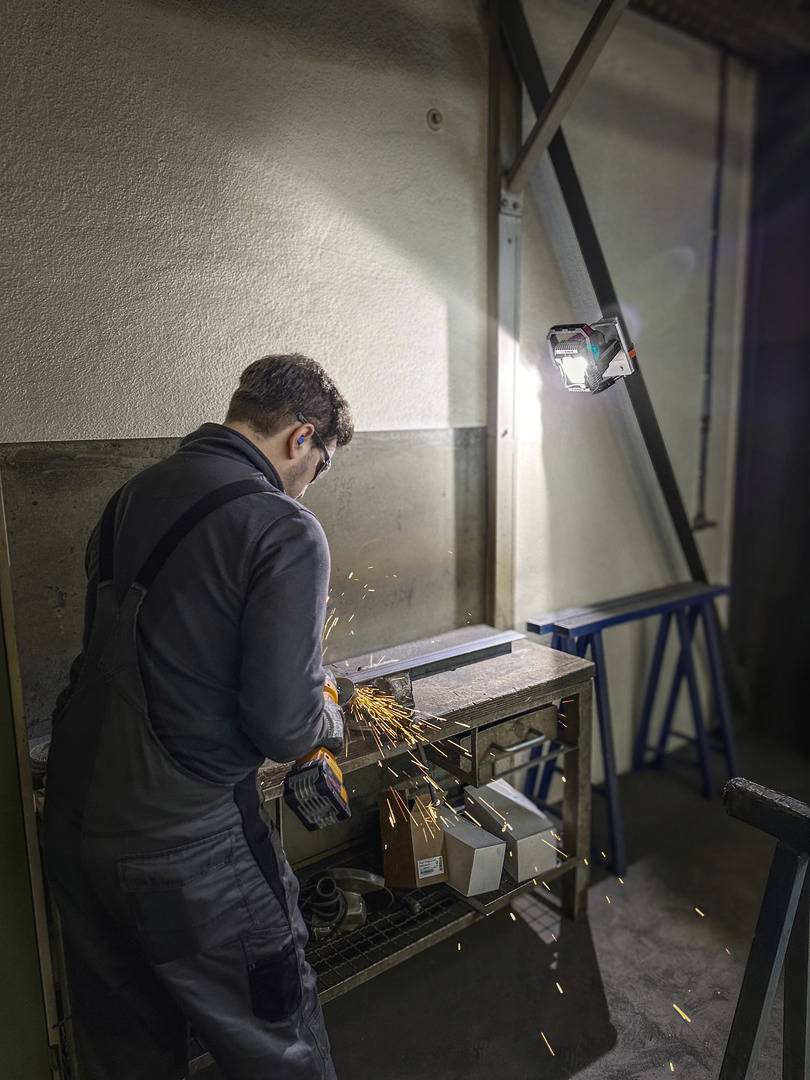 A worker in dark workwear grinds metal on a workbench. Sparks fly as a lamp illuminates the area.