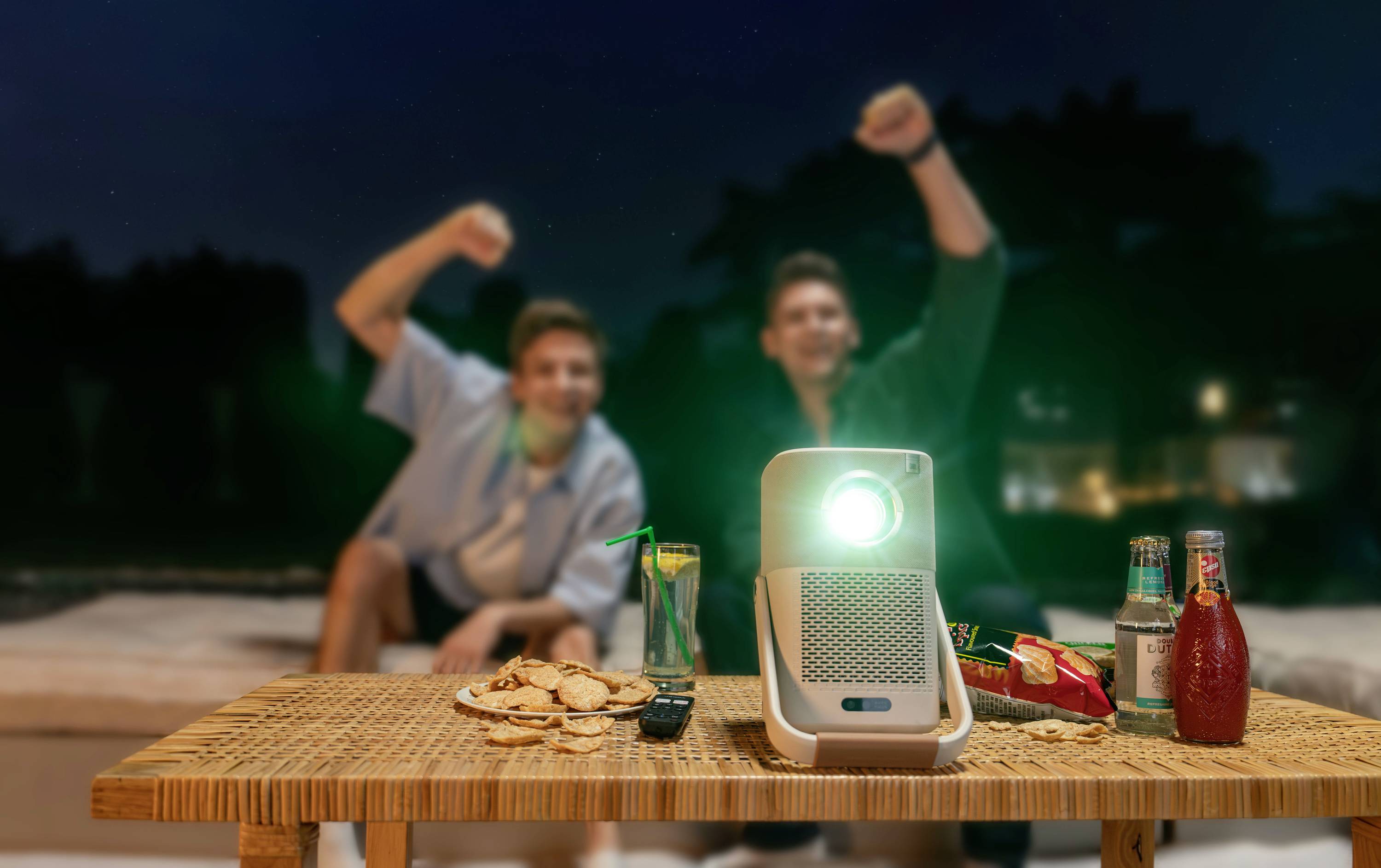 Two people are cheering on an outdoor sofa, with a projector glowing on a table, surrounded by drinks and snacks.