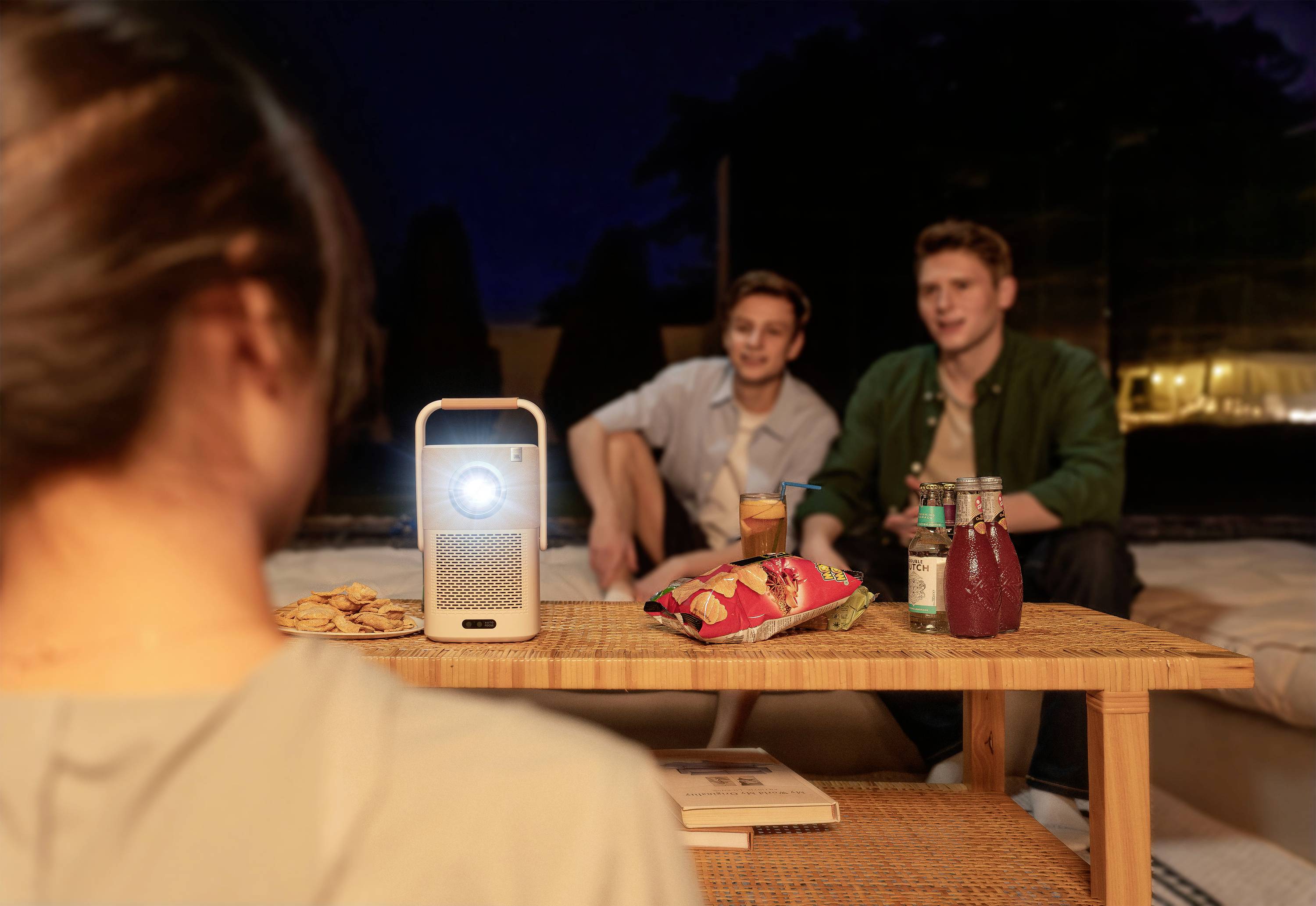A projector displays an image in a nighttime outdoor scene. Two people are seated, with drinks and snacks on a table.