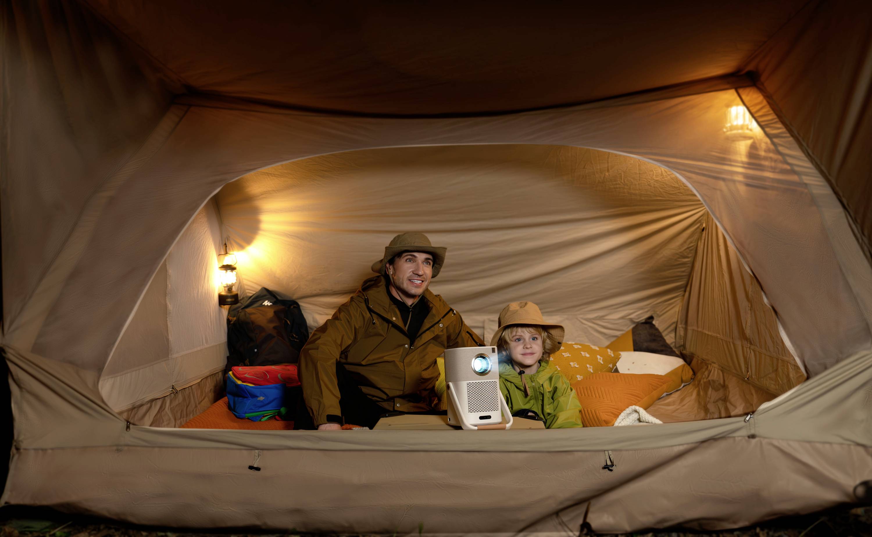 An adult and a child are sitting in an illuminated tent, looking at a projector and appearing relaxed and content.