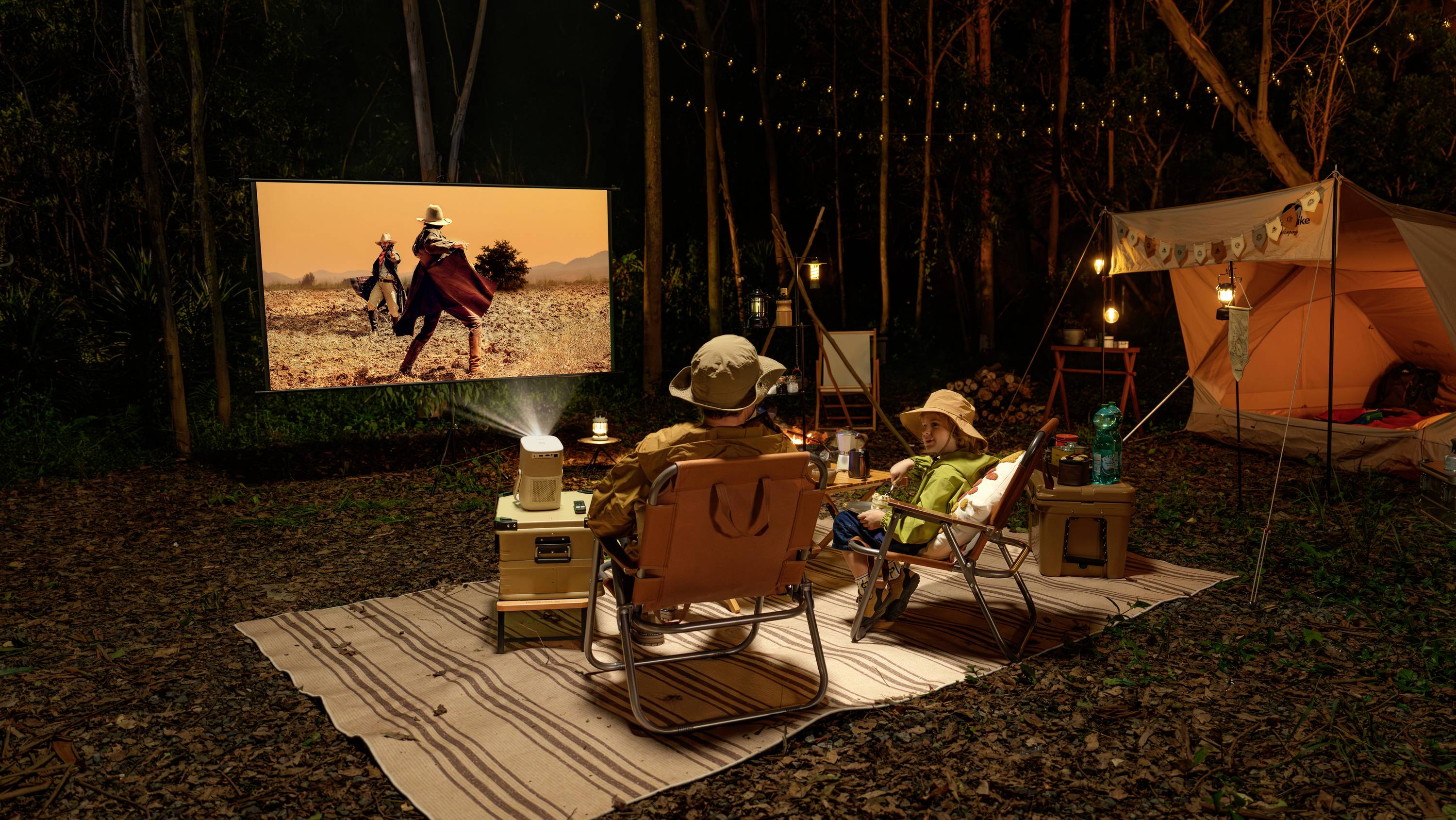 Two people are sitting on loungers in the woods in the evening, watching a film projected onto a screen by a projector.