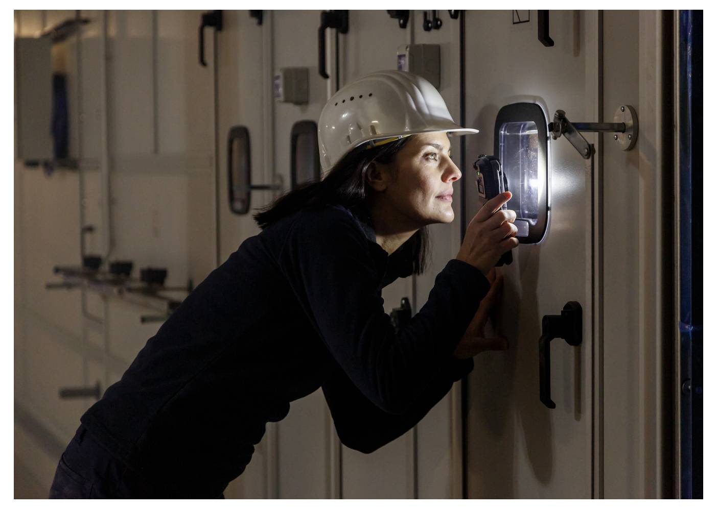 Woman in a hard hat inspects a door panel with a flashlight in a dim industrial hallway.