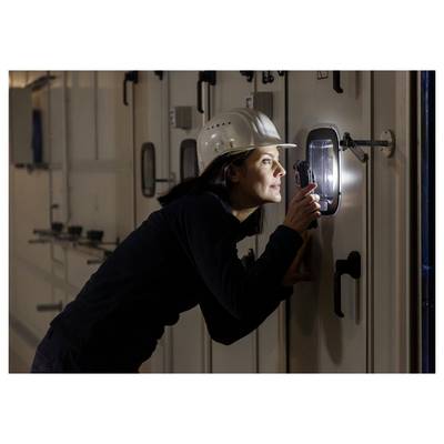 Woman in a hard hat inspects a door panel with a flashlight in a dim industrial hallway.