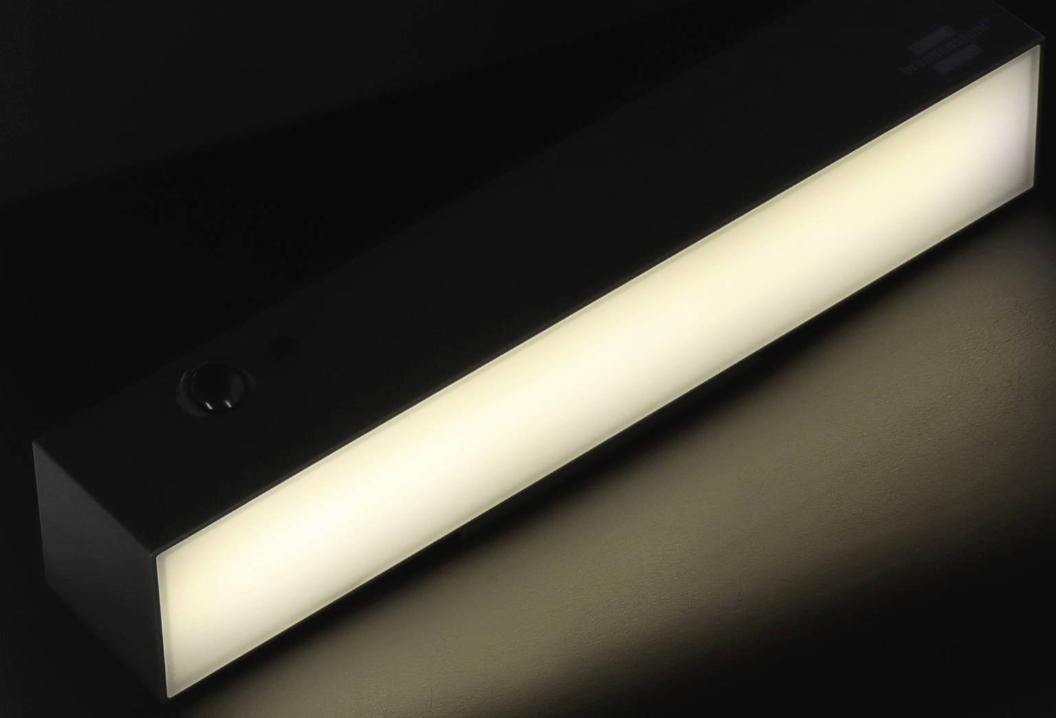 A rectangular LED light bar illuminates in a dark room. The light radiates evenly and lights up the surrounding area.