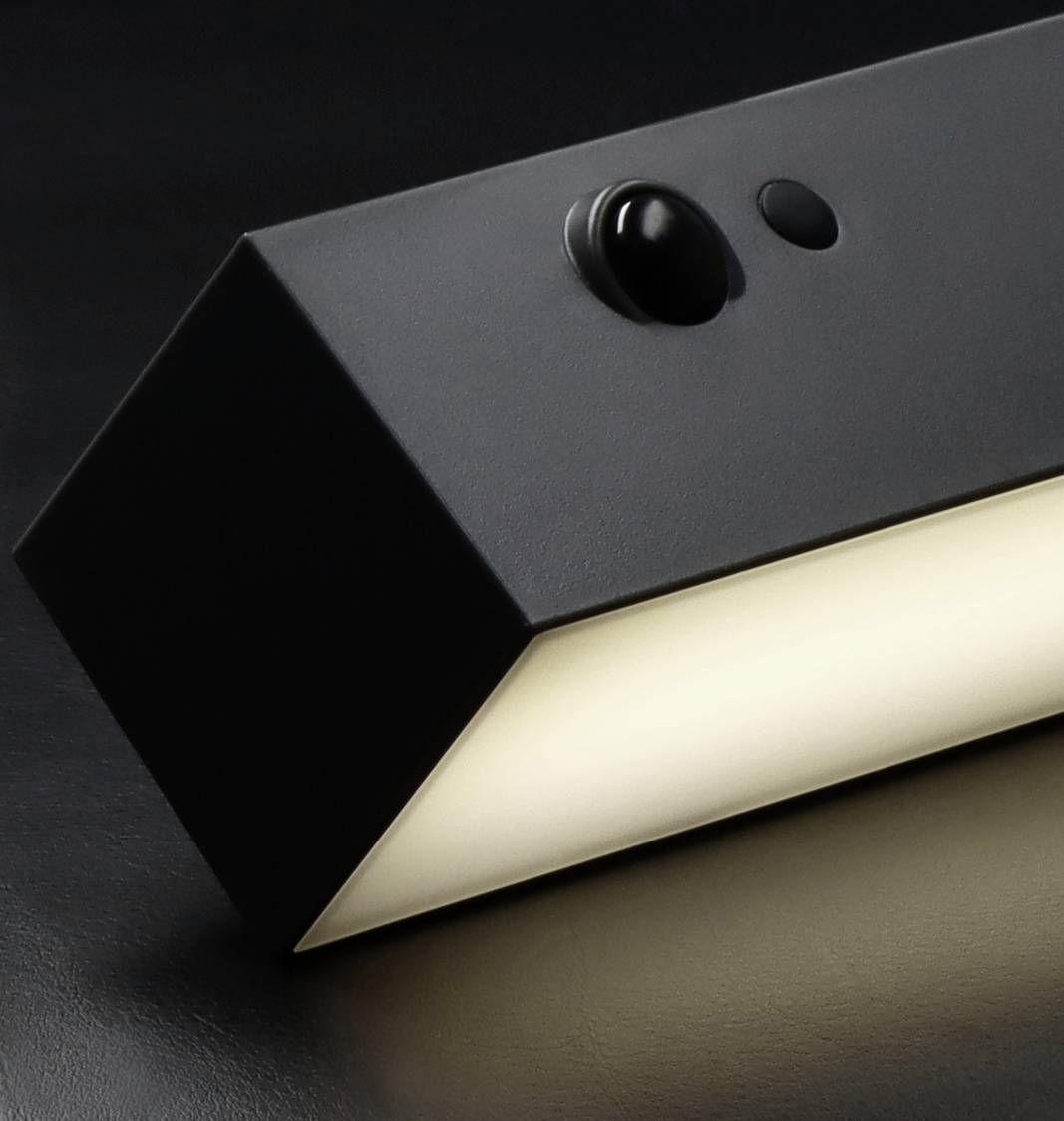 Black, rectangular wall light on a dark background, illuminating brightly from below with a motion sensor at the top.