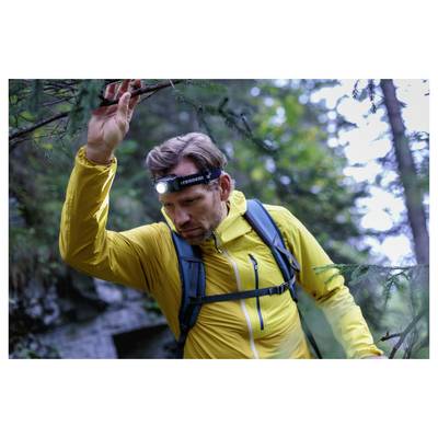 Hiker in a yellow jacket with a headlamp raises one arm while walking through a forest.
