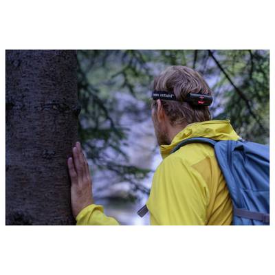 A hiker in a yellow jacket and blue backpack presses a hand against a tree while wearing a headlamp in a wooded area.