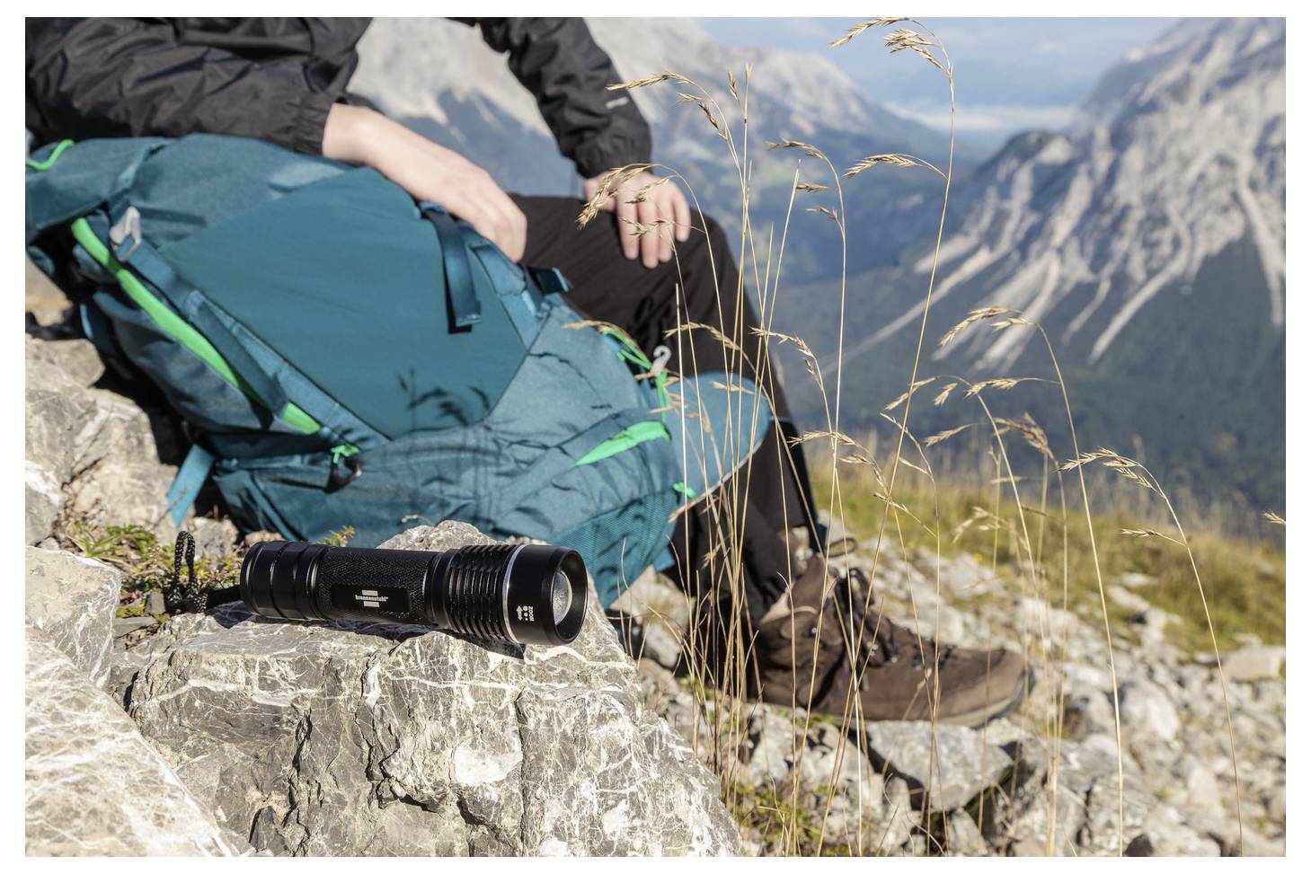 Hiker resting on a rocky mountain ledge with a teal backpack, boots, and a camera lens in front; alpine peaks in the background.