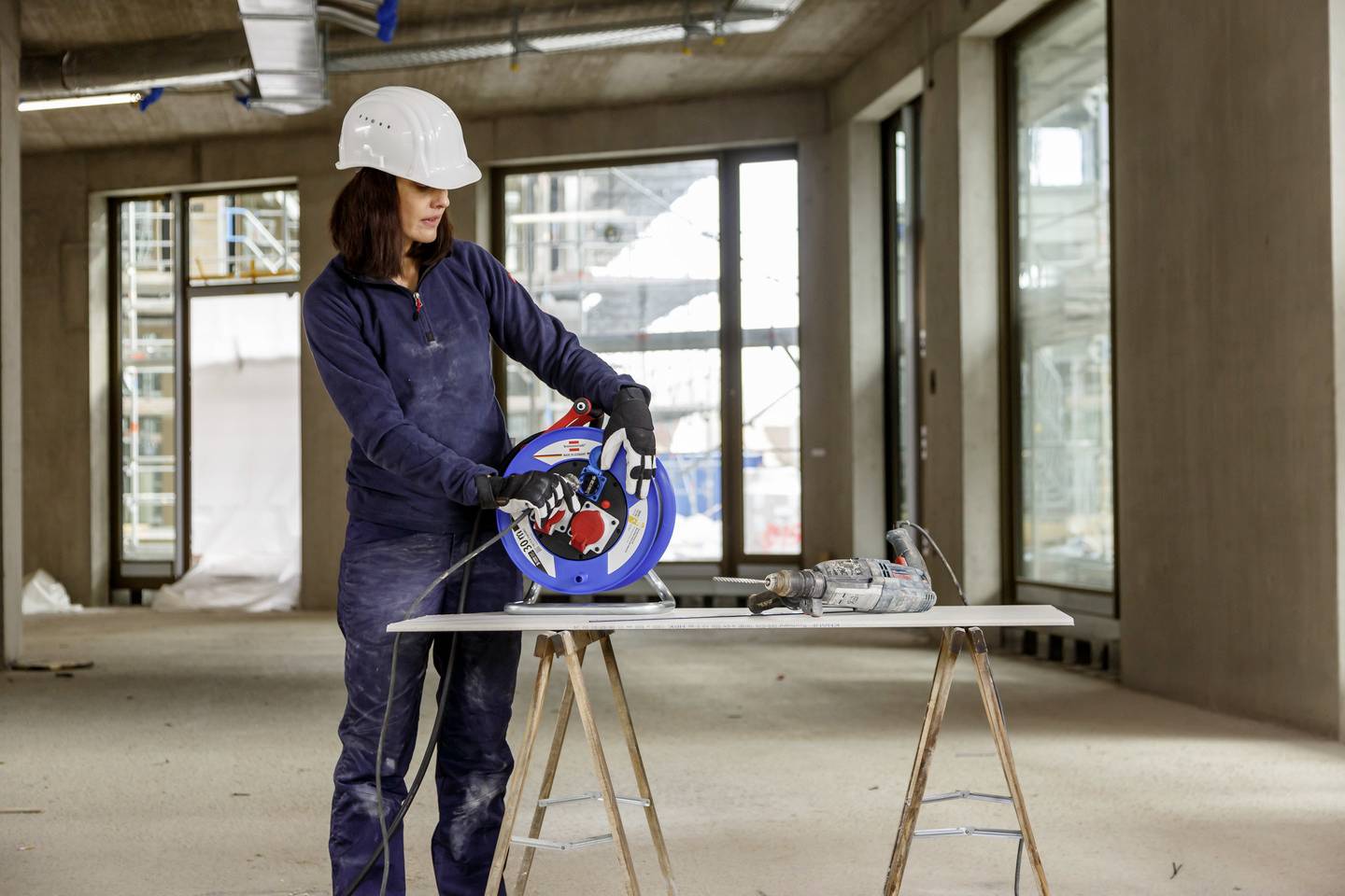 A female construction worker in protective clothing stands in an unfinished room, holding a cable reel and preparing a drill on a table.