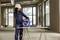 A female construction worker in protective clothing stands in an unfinished room, holding a cable reel and preparing a drill on a table.
