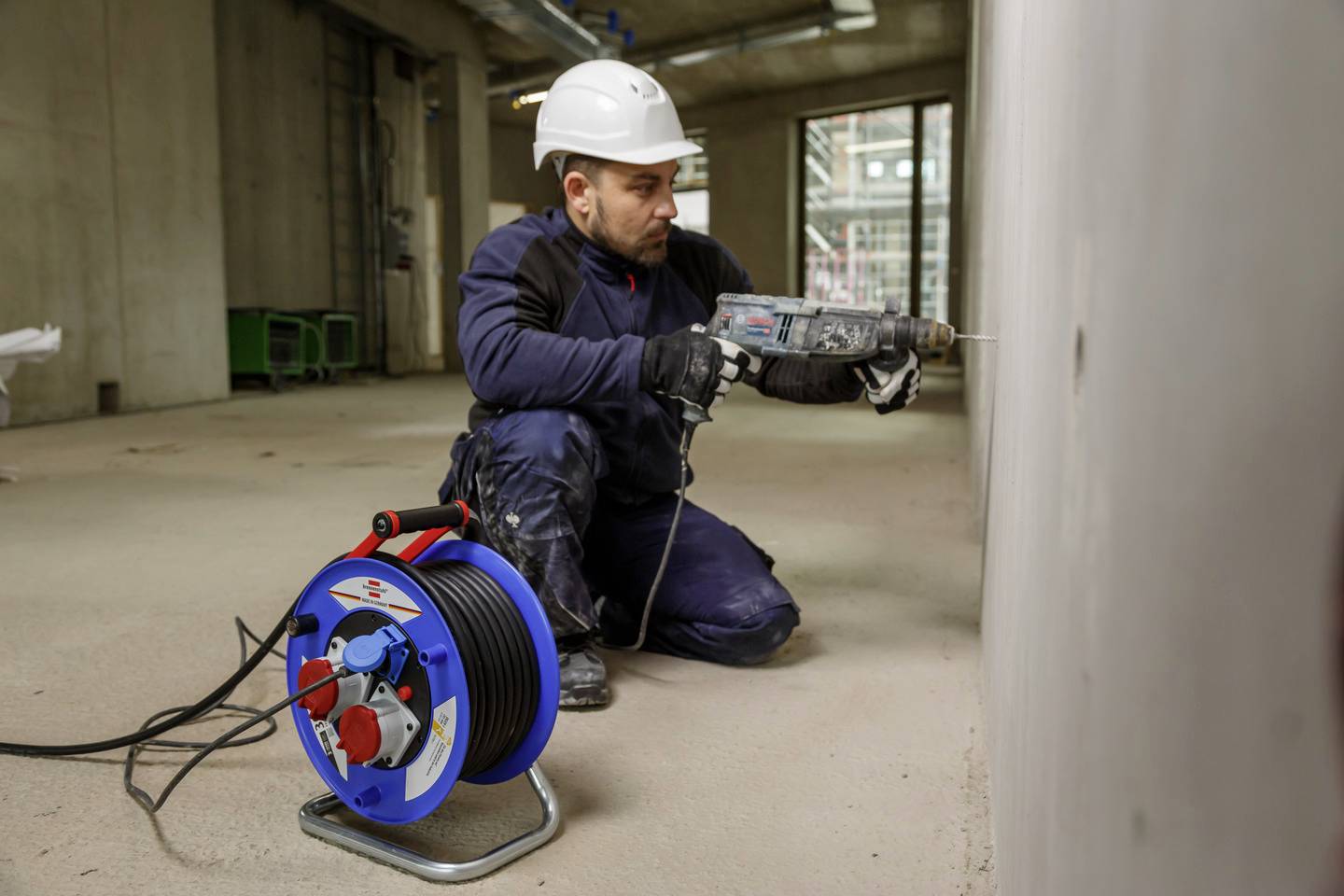A construction worker is wearing a white hard hat and drilling a hole in a wall on a building site. A cable drum lies in the foreground.