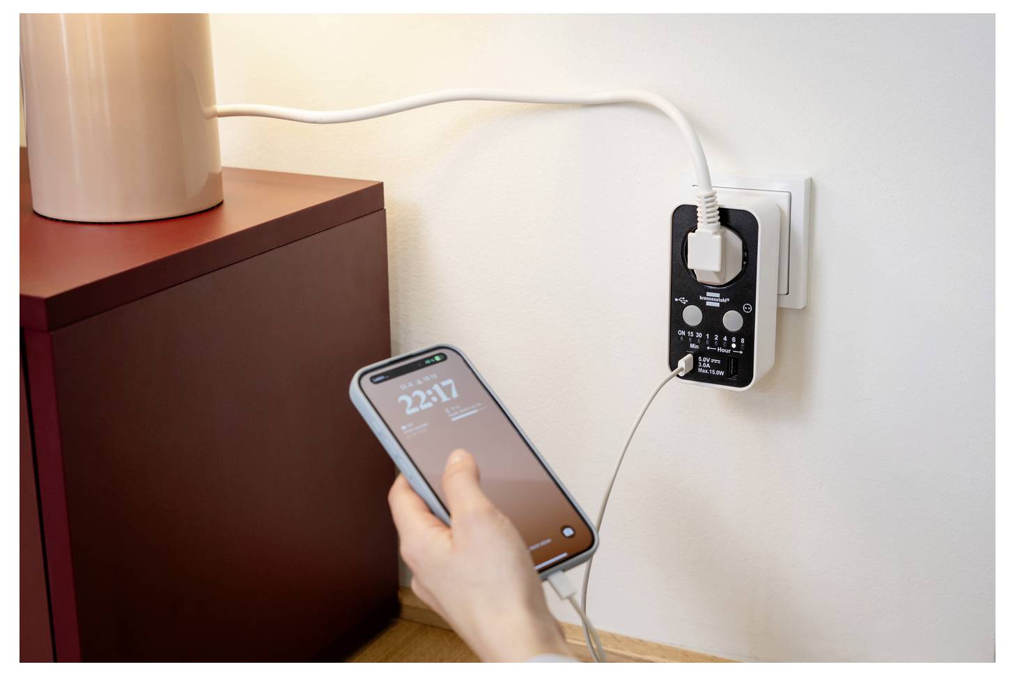 Hand holding a smartphone plugged into a wall charger beside a cabinet, showing the phone charging from an outlet adapter.<br>