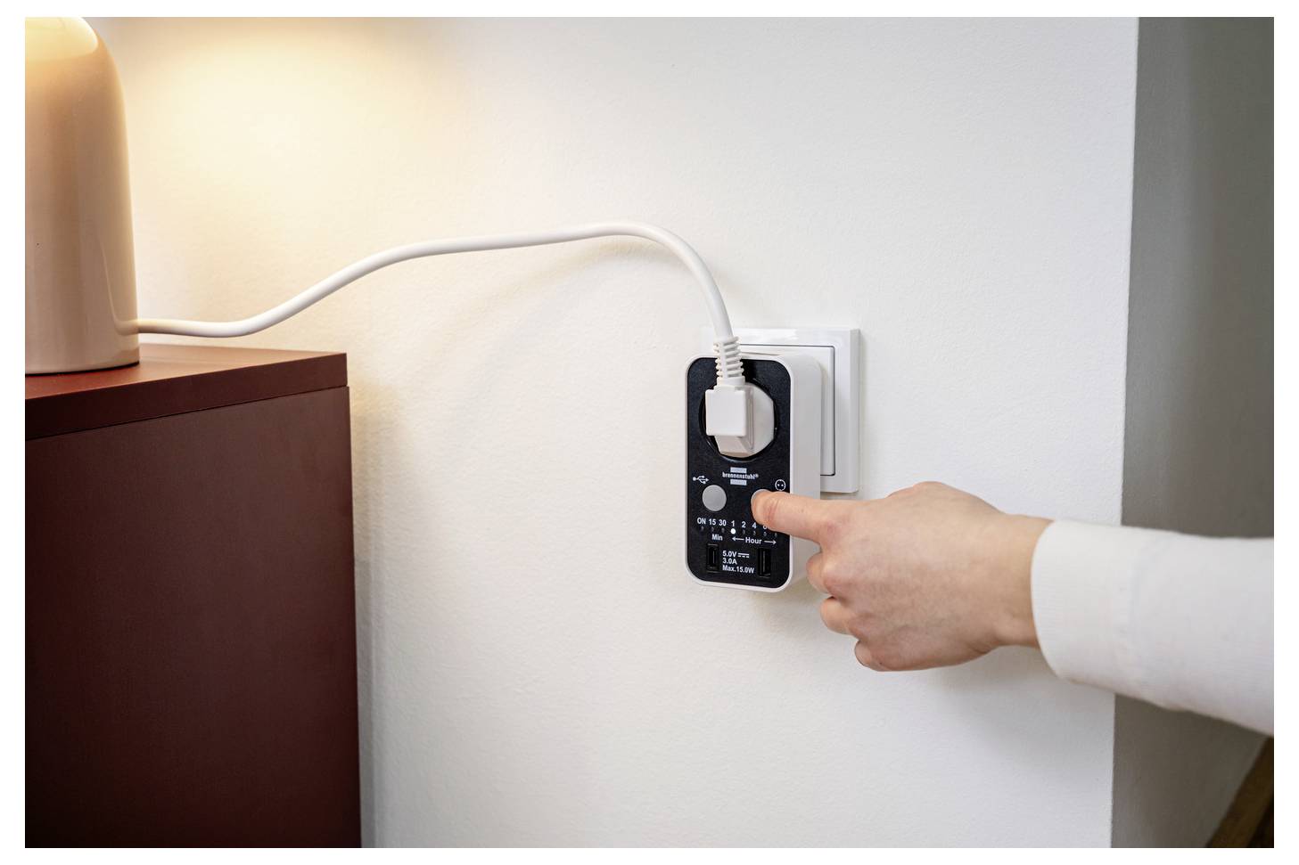 Hand plugging a lamp into a wall outlet with an inline power meter or smart plug, showing electricity usage control.