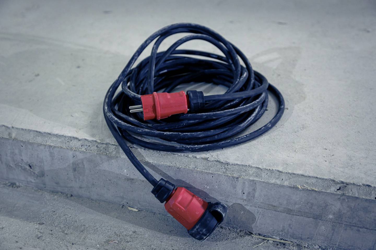 A long, black extension lead with red plugs is lying on a concrete floor.