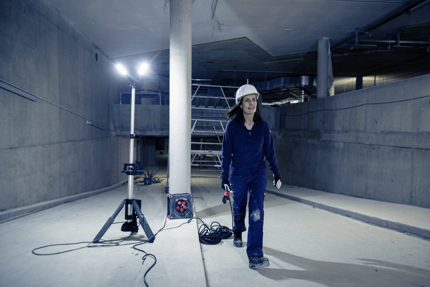 A person wearing protective clothing and a hard hat walks through a construction site with concrete walls, illuminated by a work light. Scaffolding can be seen in the background.