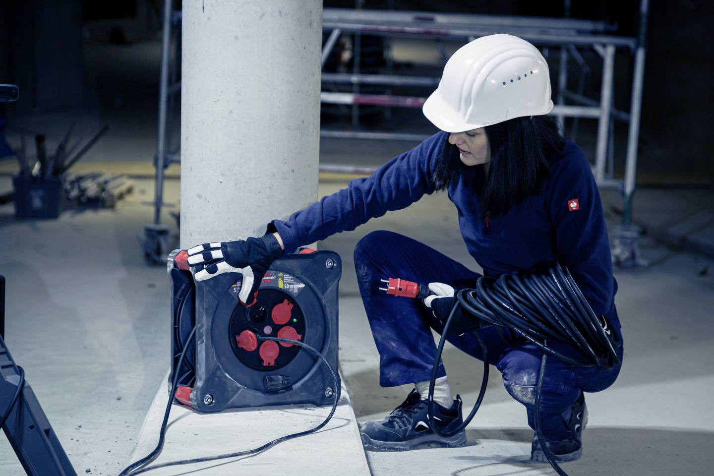 A person in work attire and a hard hat is kneeling beside a cable reel on a construction site and checking a cable.