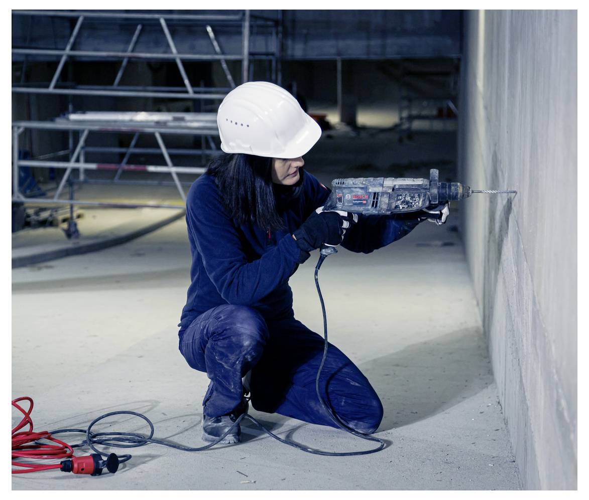 Construction worker in hard hat kneels indoors using a power drill to bore into a concrete wall, with cords on the floor and scaffolding behind.<br>