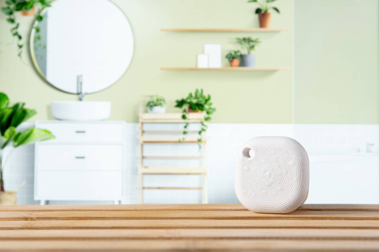 A white, square air freshener sits on a wooden shelf in a modern, bright bathroom with plants and decorations.