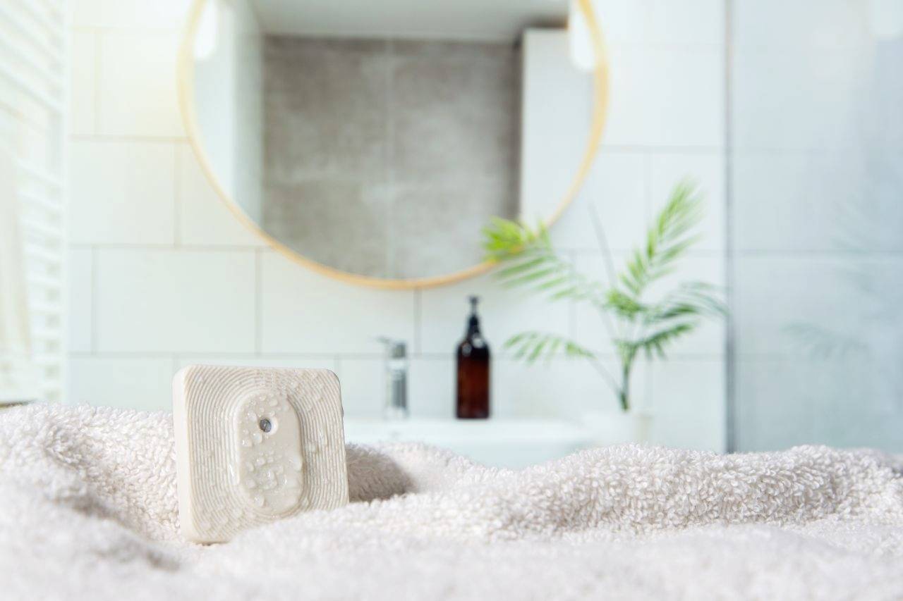 A bar of soap rests on a towel in a bathroom. In the background, a round mirror and plants can be seen.