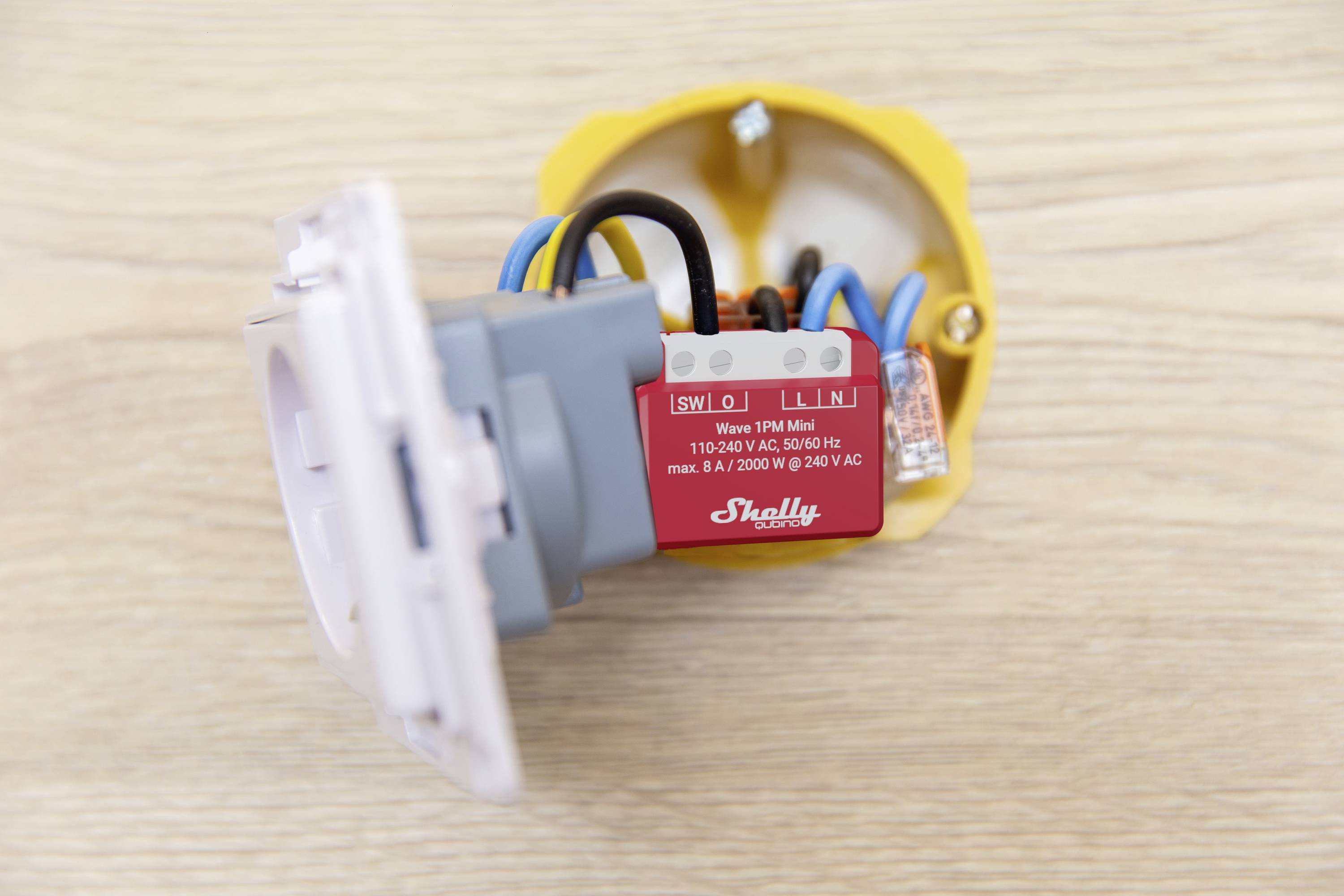 A red Shelly Wi-Fi relay component is connected to several coloured cables in a yellow wall socket on a light wooden floor.