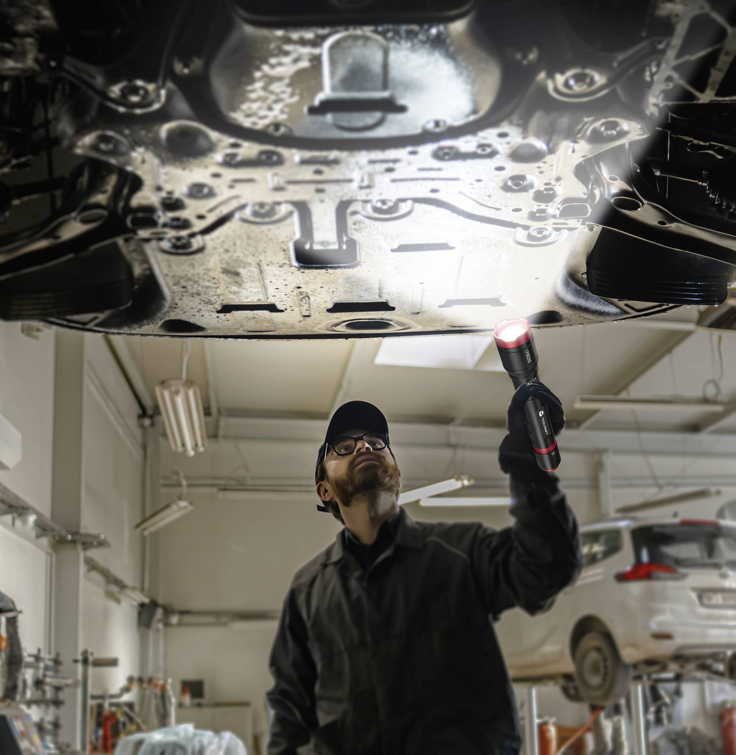 A mechanic inspects the underside of a car in a workshop with a torch, looking to identify potential issues.