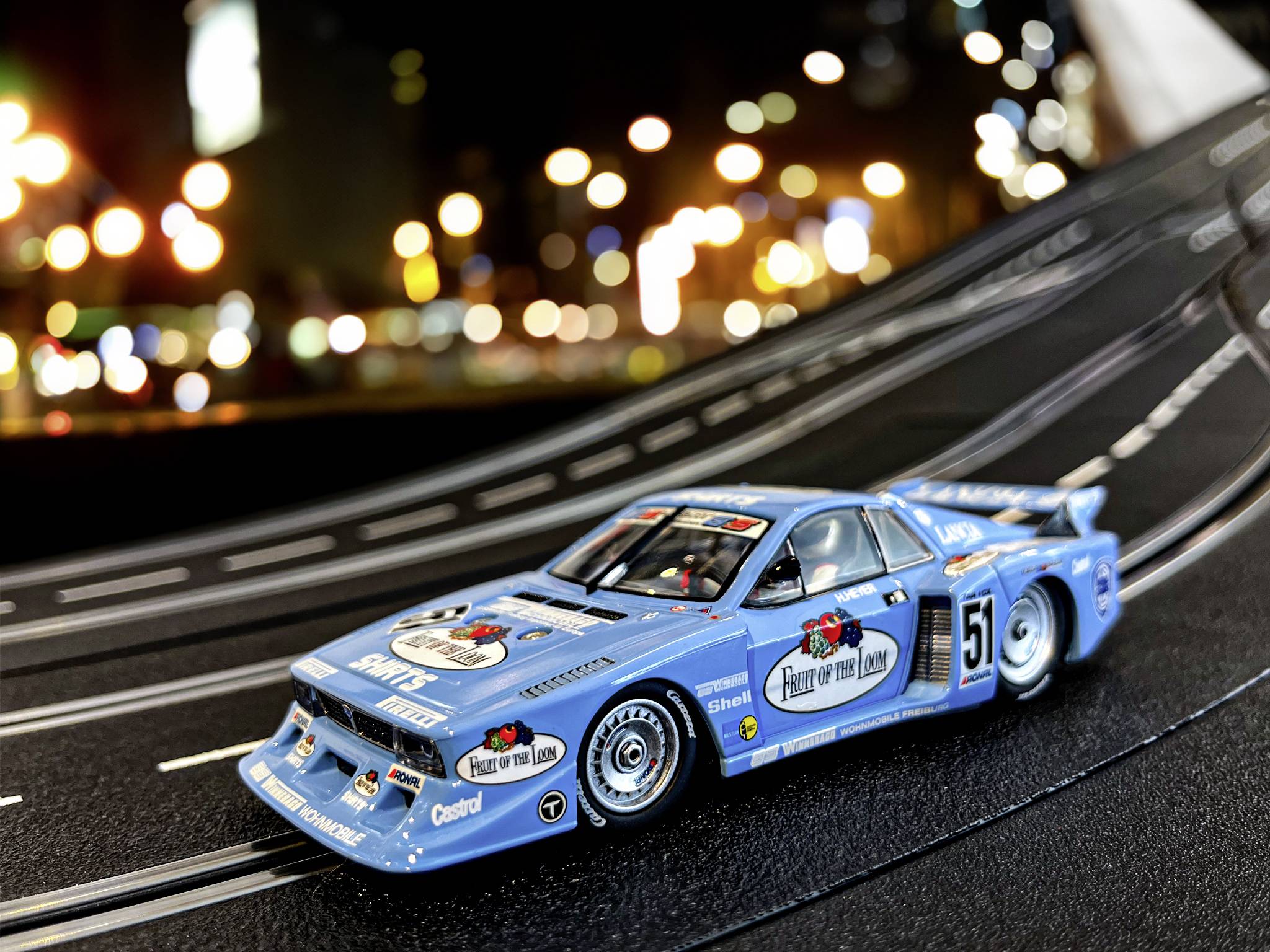 A blue model racing car with the number 51 on a racetrack at night, with an illuminated city skyline in the background.