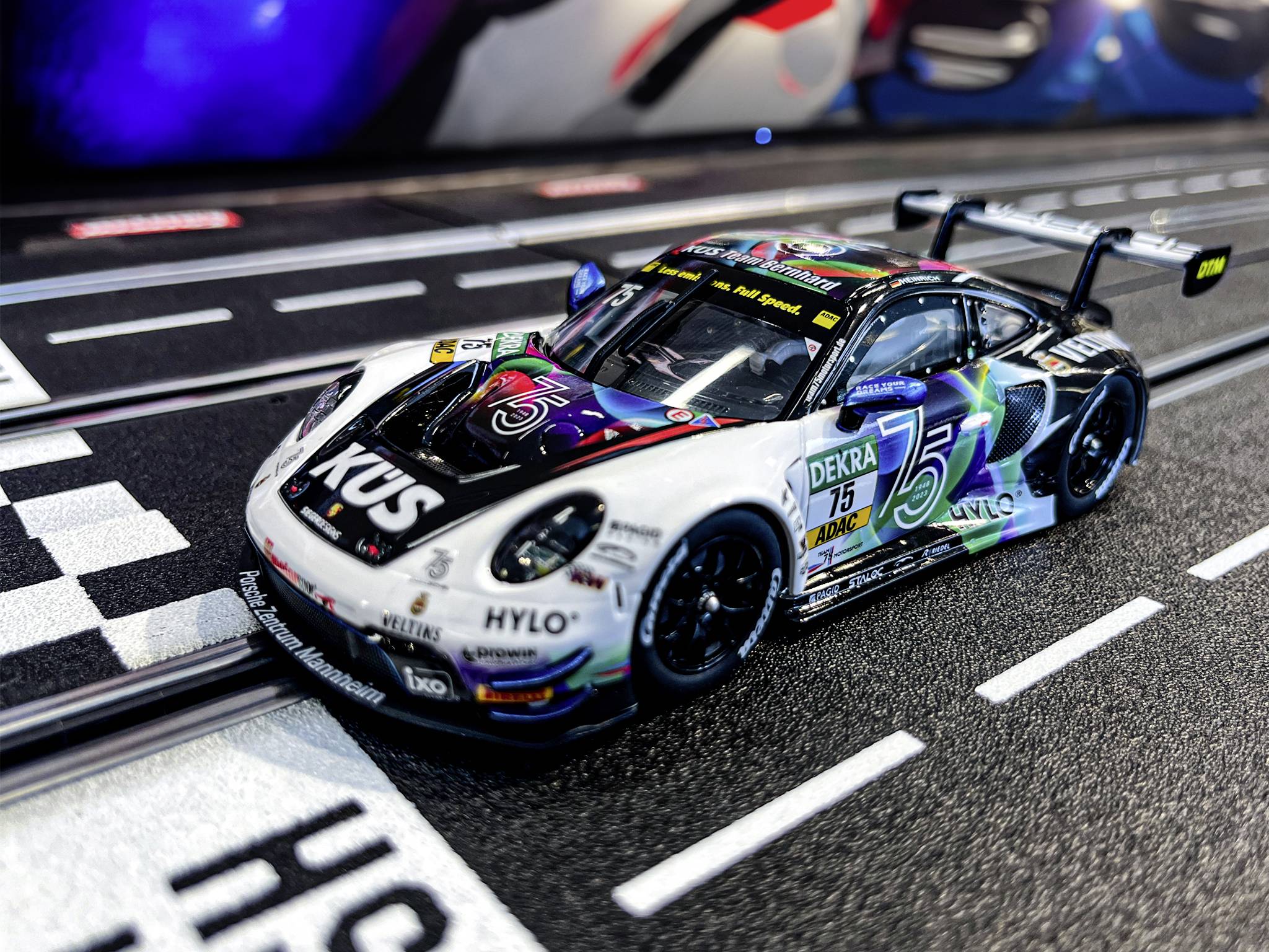 A toy racing car is positioned on a slot car racing track, ready to start. The bonnet features the 'KÜS' logo.