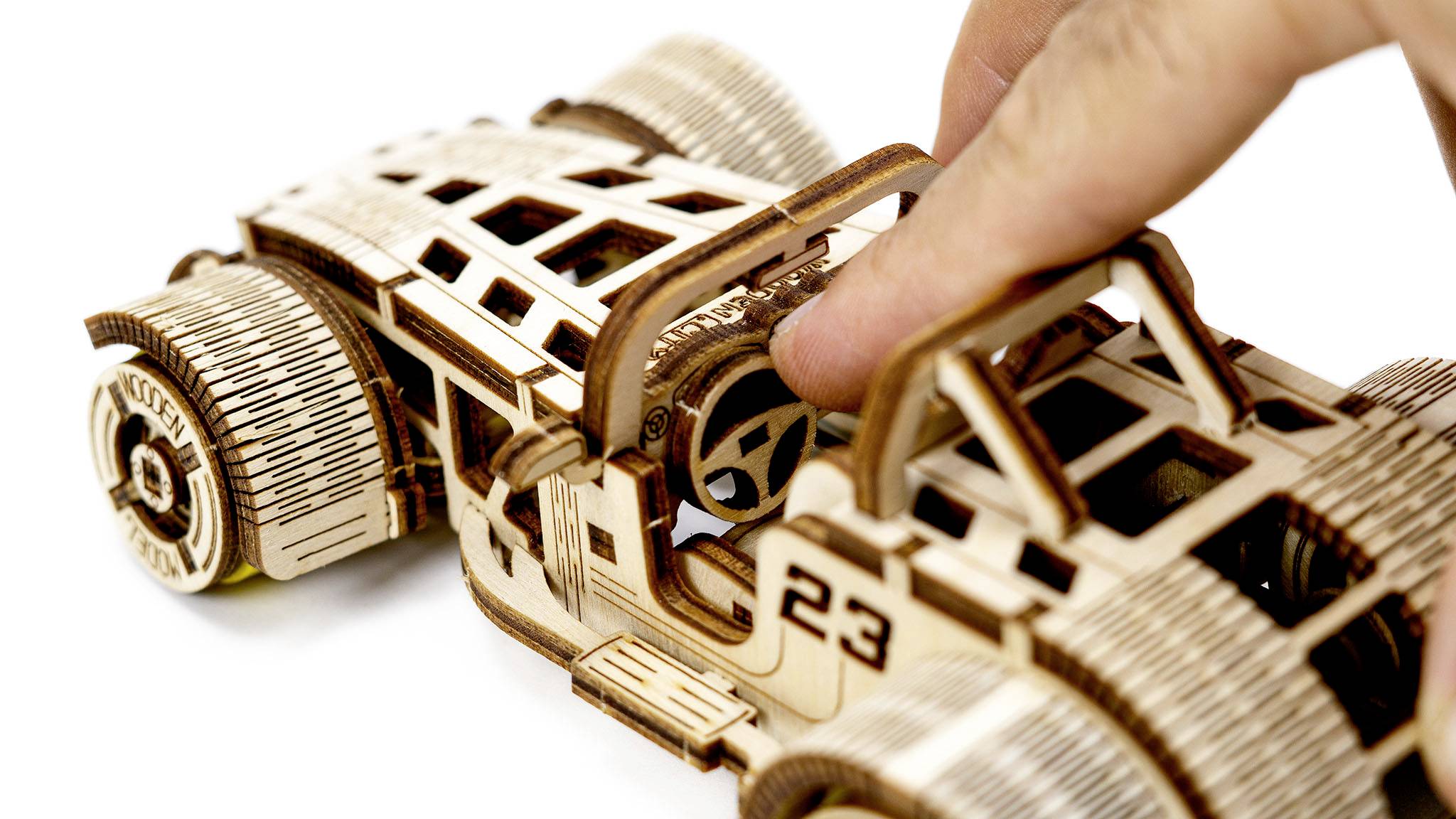 A hand touches a detailed, wooden model car with visible gears and a start number '23' on its side.
