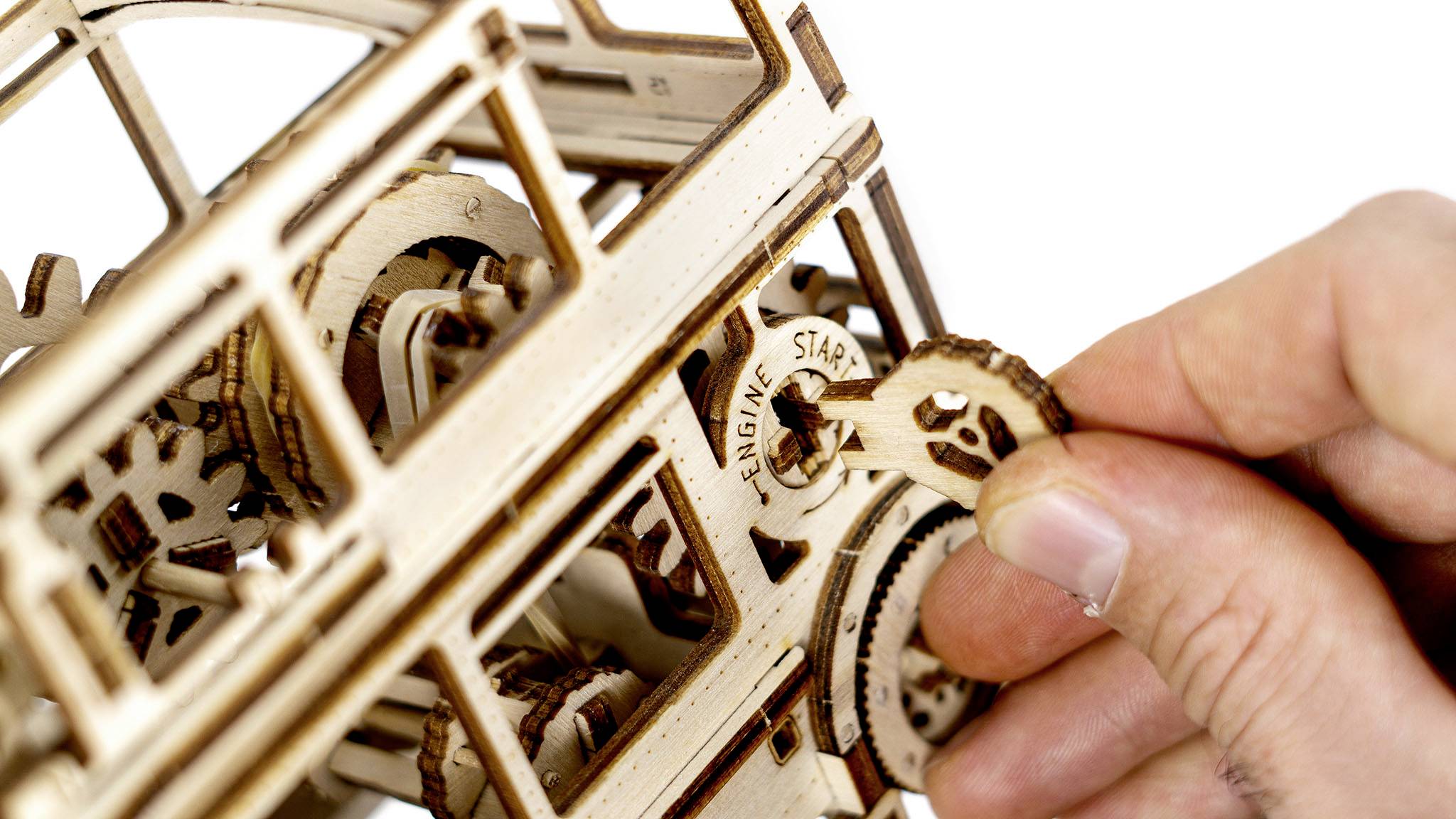 A hand turns a wooden key in a mechanical wooden model car, revealing gears and the word 'ENGINE START'.