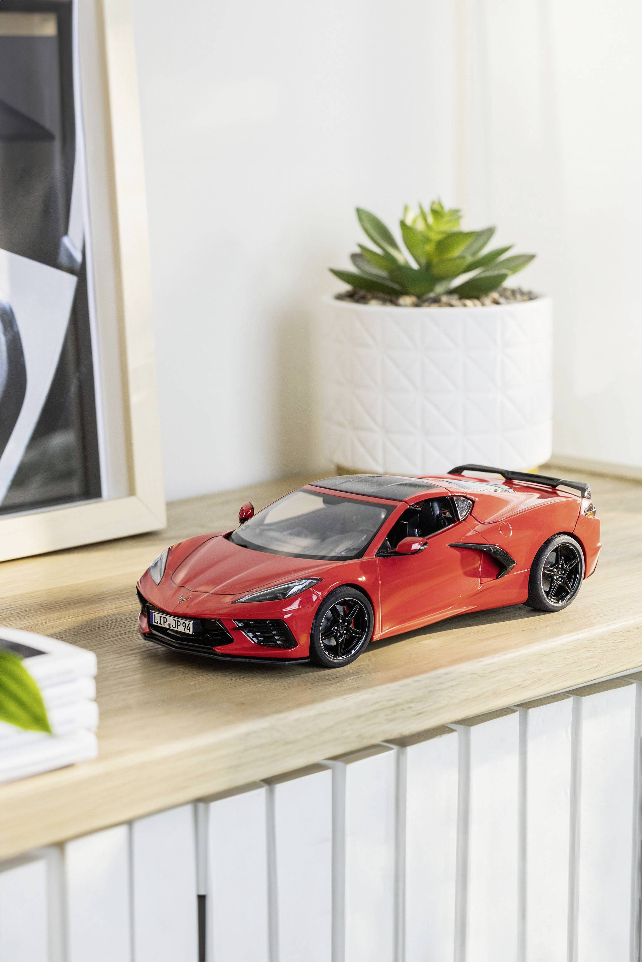 A red model car sits on a shelf, next to a plant in a white plant pot.
