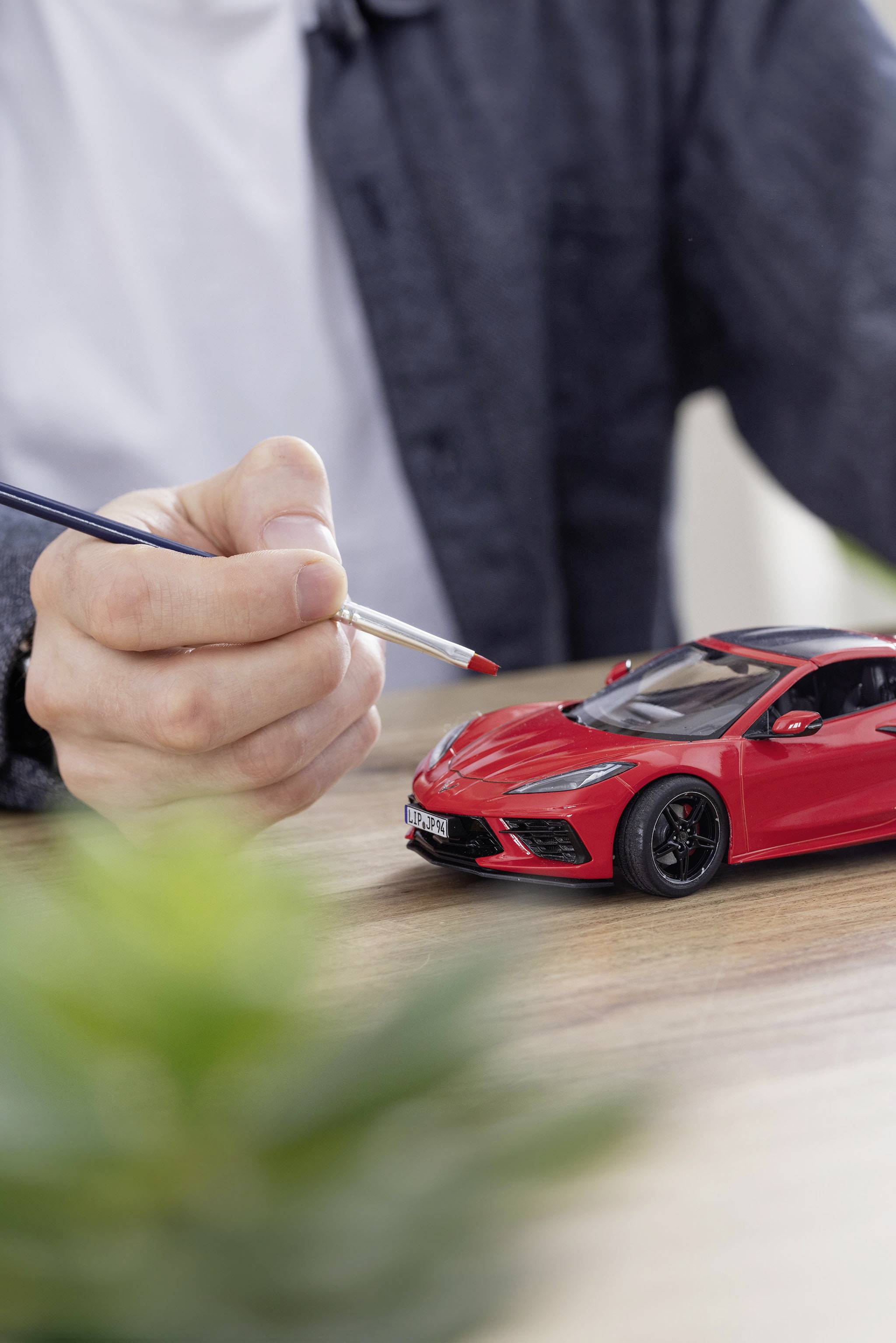 A person is painting a red model car with a brush on a wooden table. A green plant is blurred in the foreground.