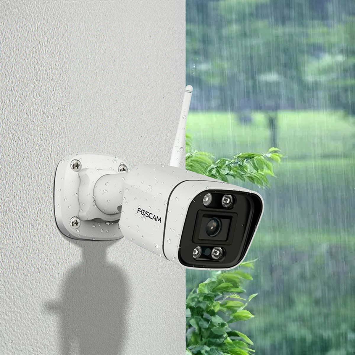A surveillance camera is mounted on an exterior wall. The lens is pointing to the right. Green leaves and a rainy sky can be seen in the background.
