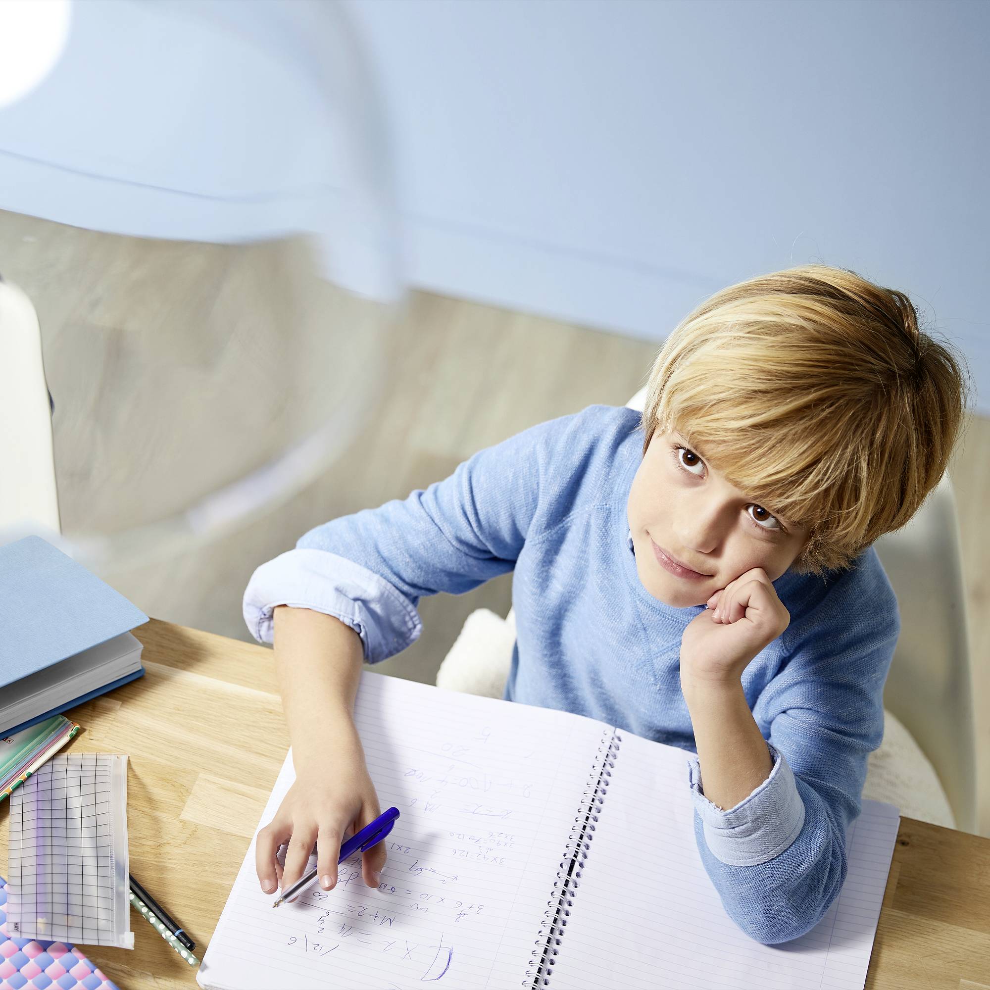 A boy sits at a table, deep in thought. In front of him lies an open notebook with some notes and pens.