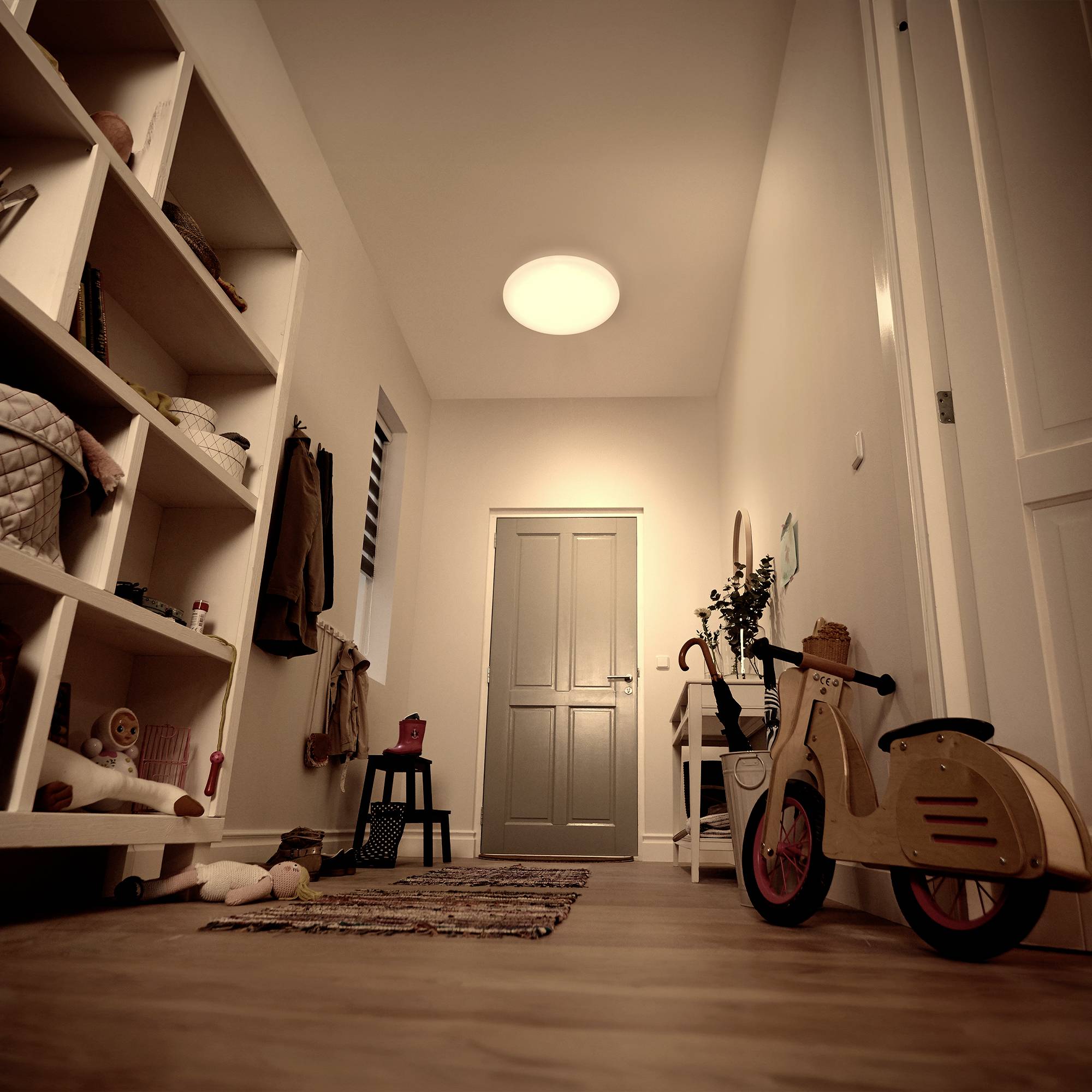 A child-decorated hallway with toys, including a wooden scooter, leads to a closed door. Right wall with shelves.
