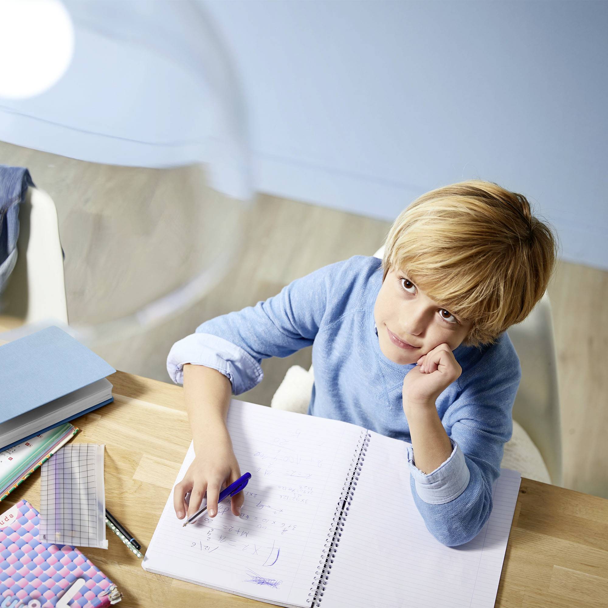 A child sits at a table, looks upwards and holds a pen over an open notebook. Books and a geometrical set square are lying on the table.