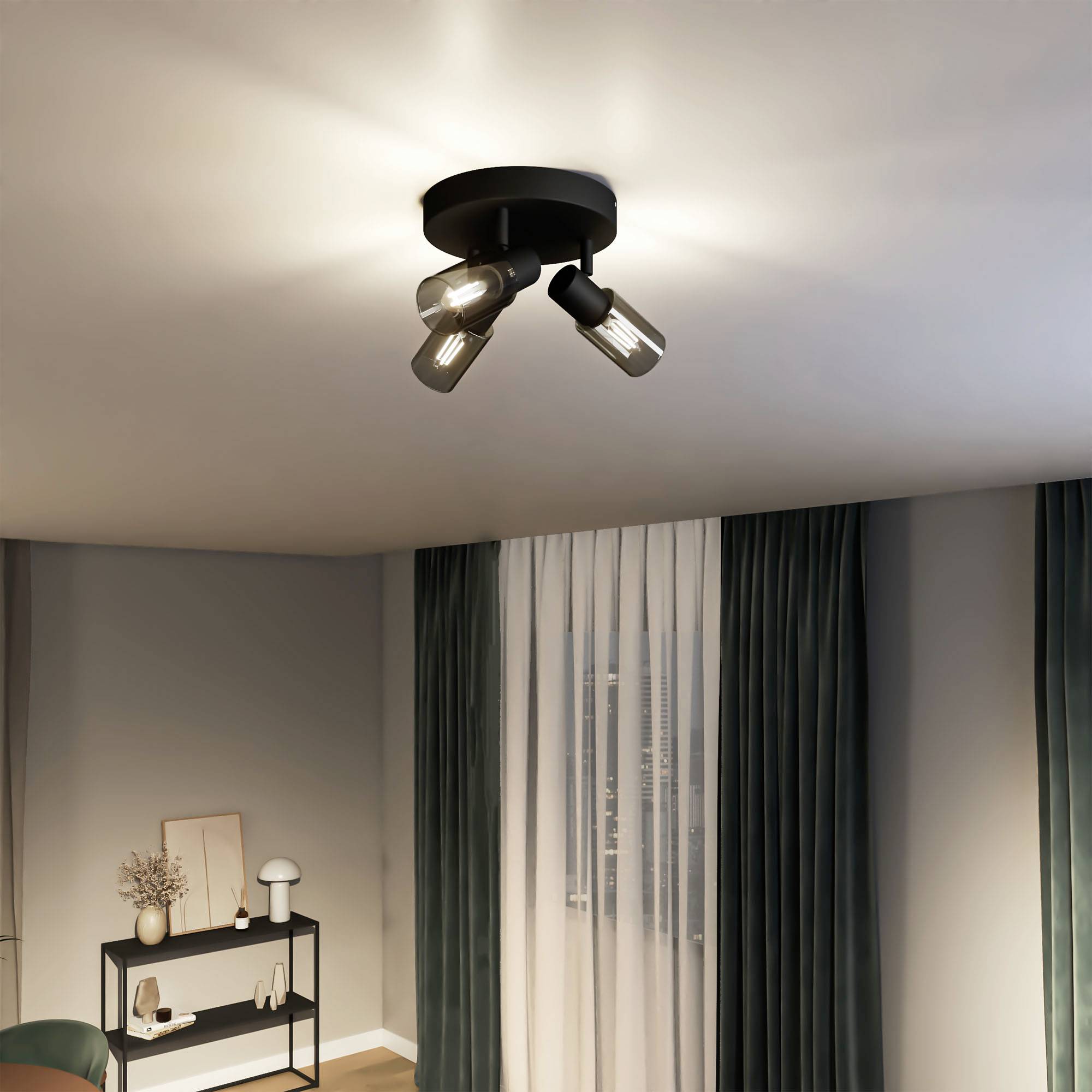 A modern ceiling spotlight with three light bulbs, illuminating a living space with green curtains and a table in the corner.