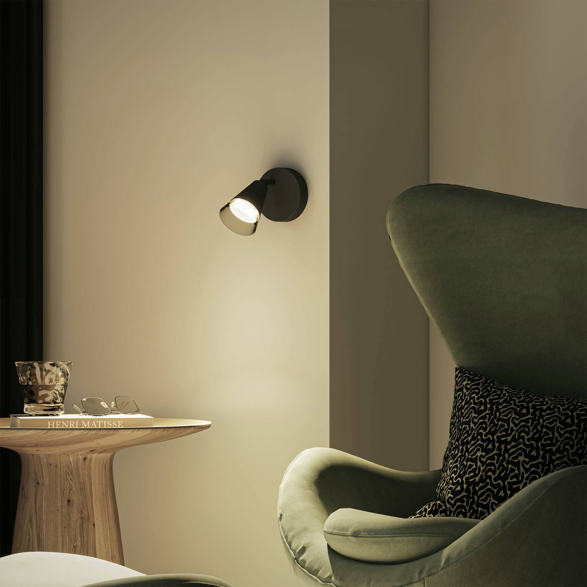 Cosy living room with a green armchair, cushions, a wooden side table with a book, and a wall lamp softly illuminating the space.