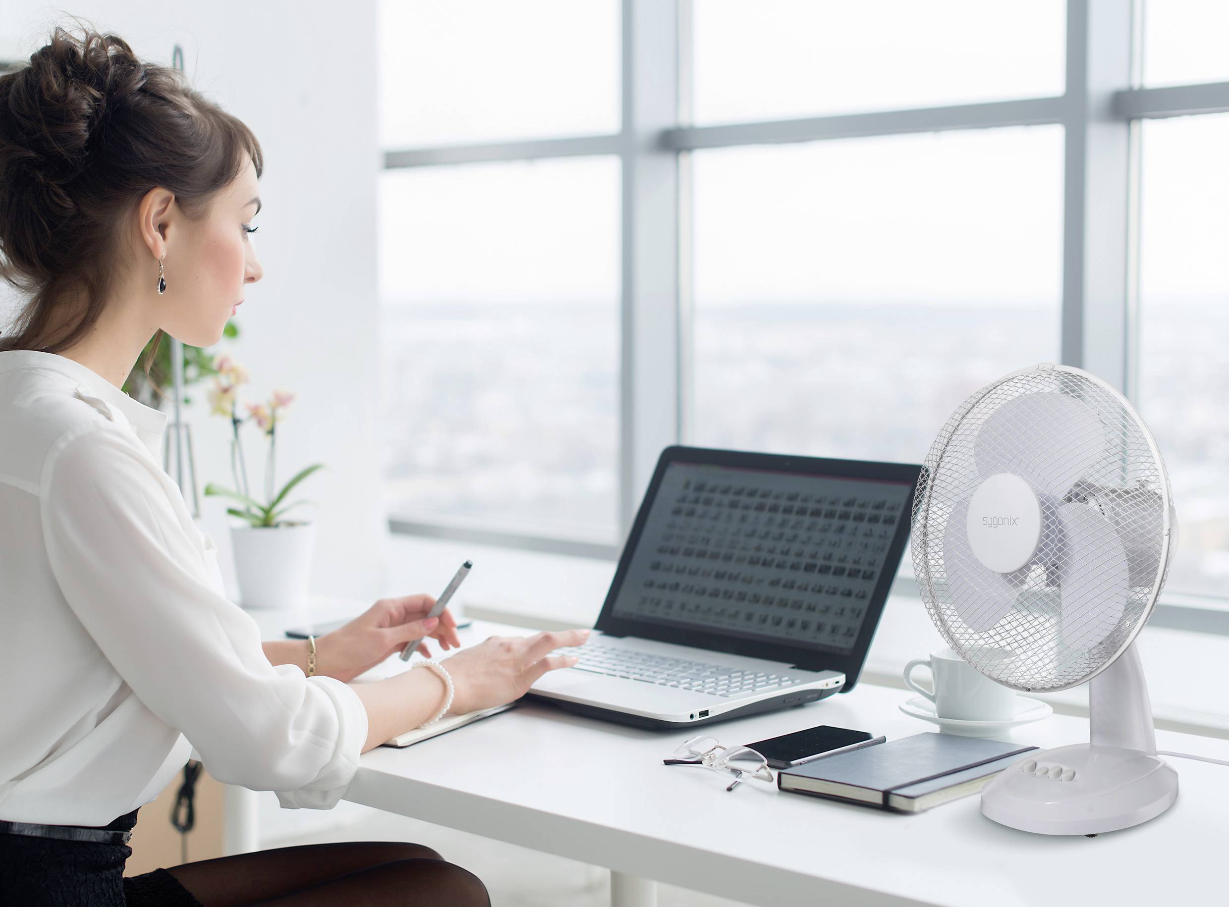 A woman is working intently on a laptop at a desk, with notebooks, glasses, and a desk fan beside her.