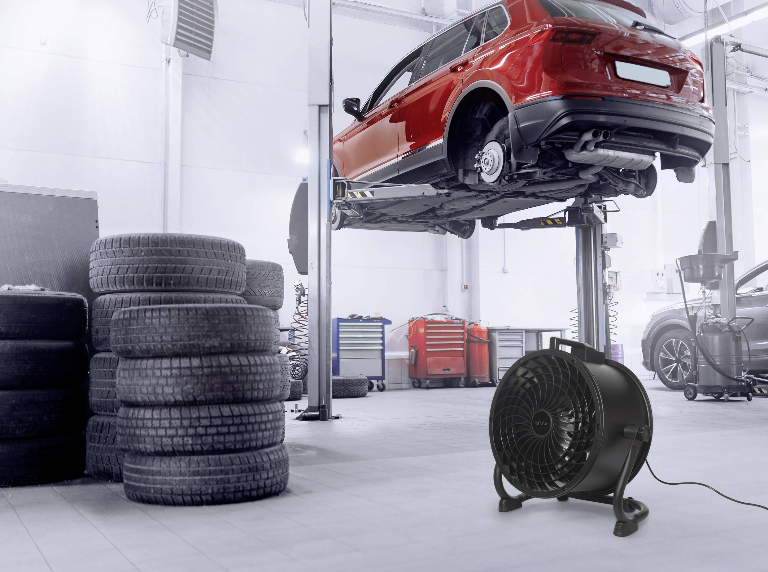 A red car is raised on a hydraulic lift in a garage workshop. Stacked tyres are on the left, with a fan in the foreground.