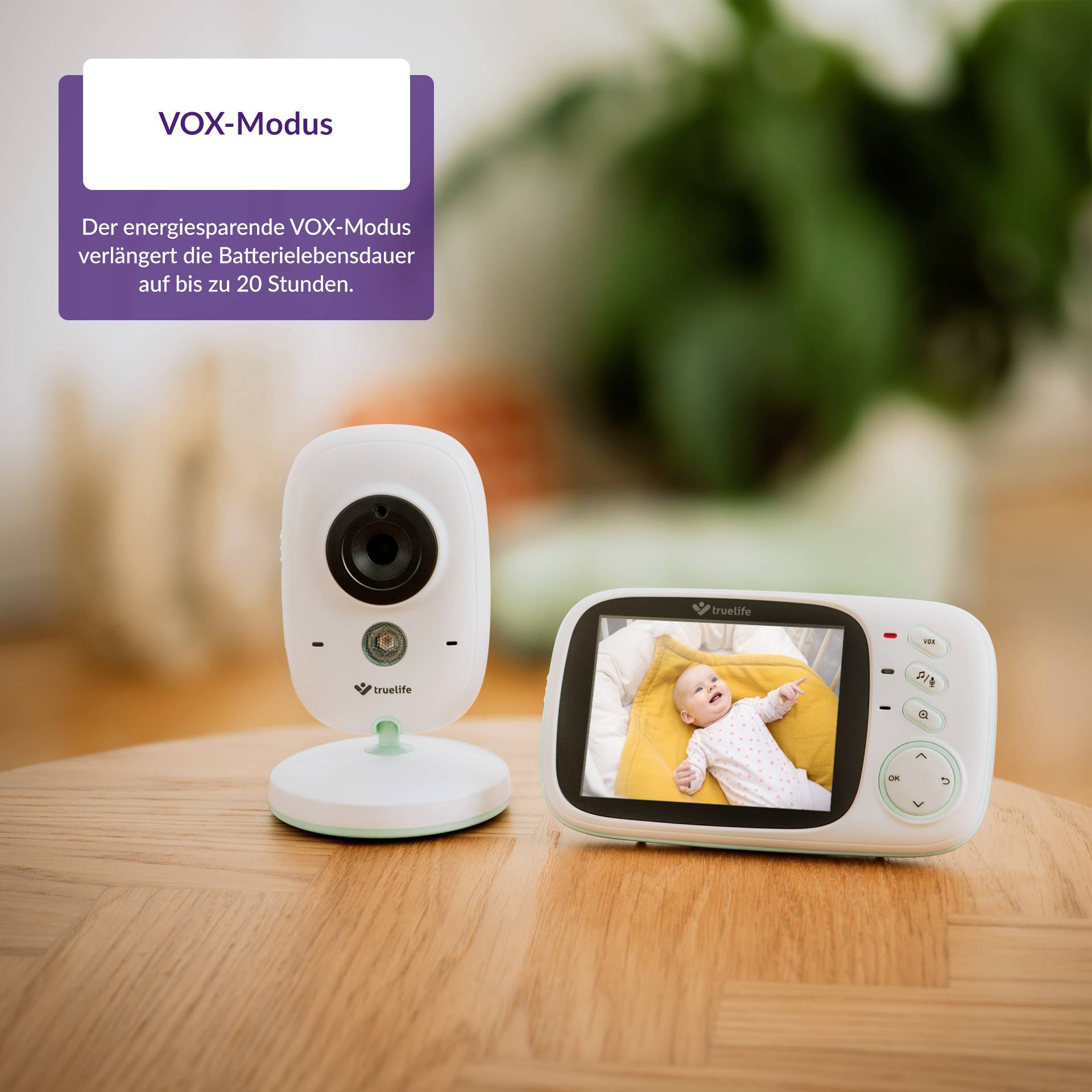 'Baby monitor on wooden table, camera on the left, monitor on the right showing baby. Text emphasises energy-saving VOX mode with up to 20 hours of battery life.'