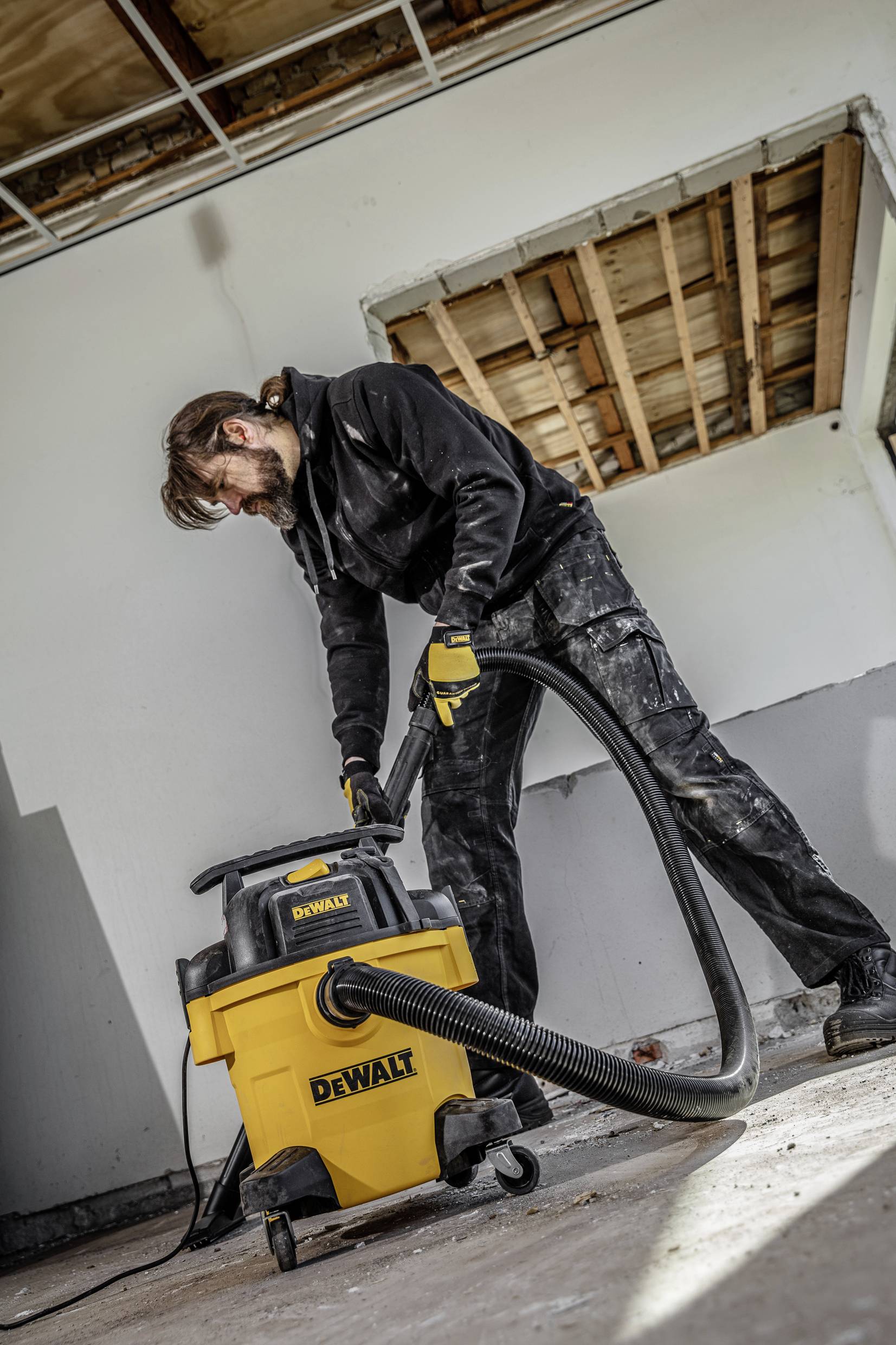 A man in work attire is operating a yellow workshop vacuum cleaner in a partially completed room. Ceiling partially exposed, floor clean.