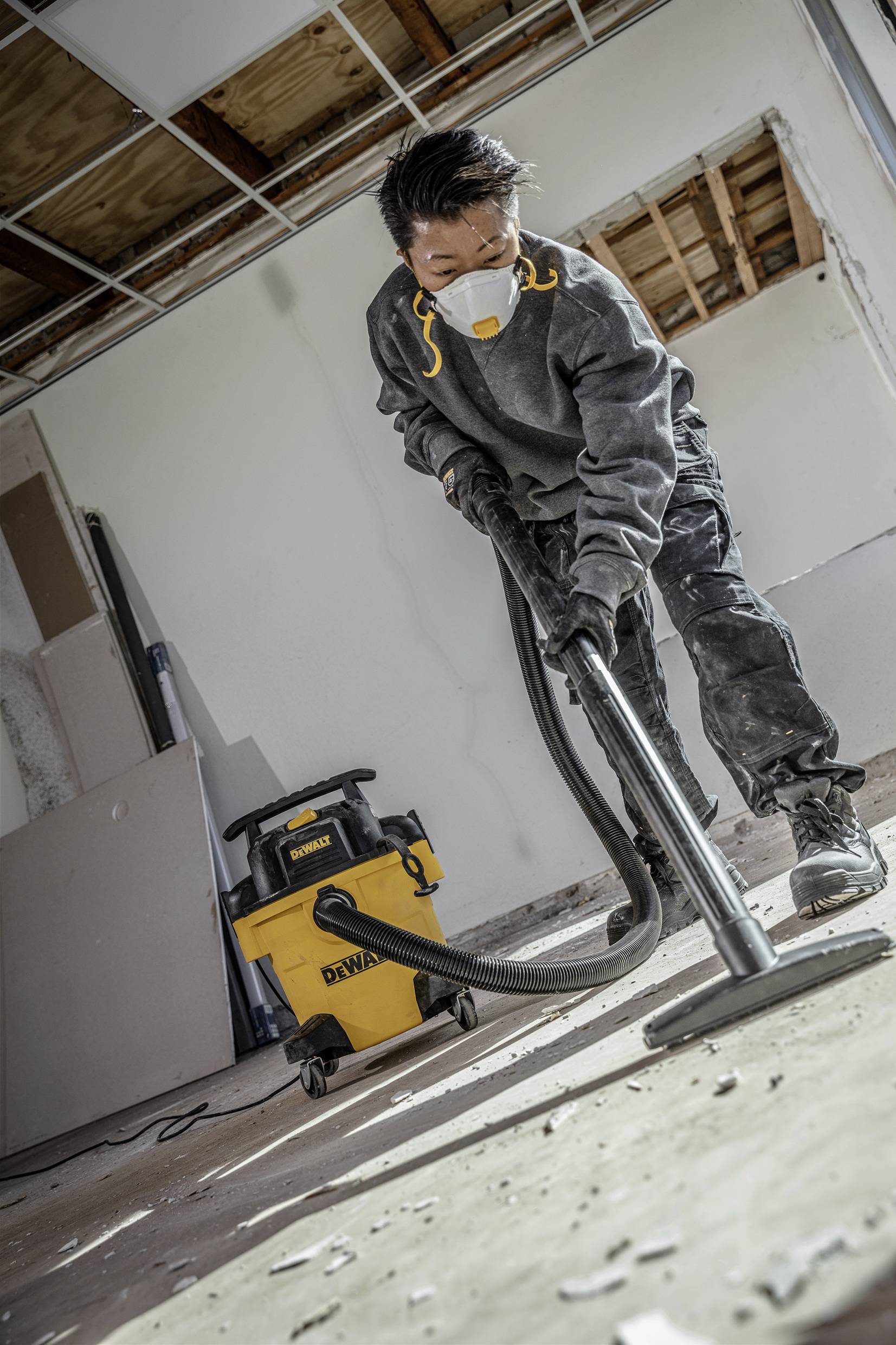 A person wearing a protective mask is vacuuming dust and debris from the floor in a room under construction. A yellow industrial vacuum cleaner stands nearby.
