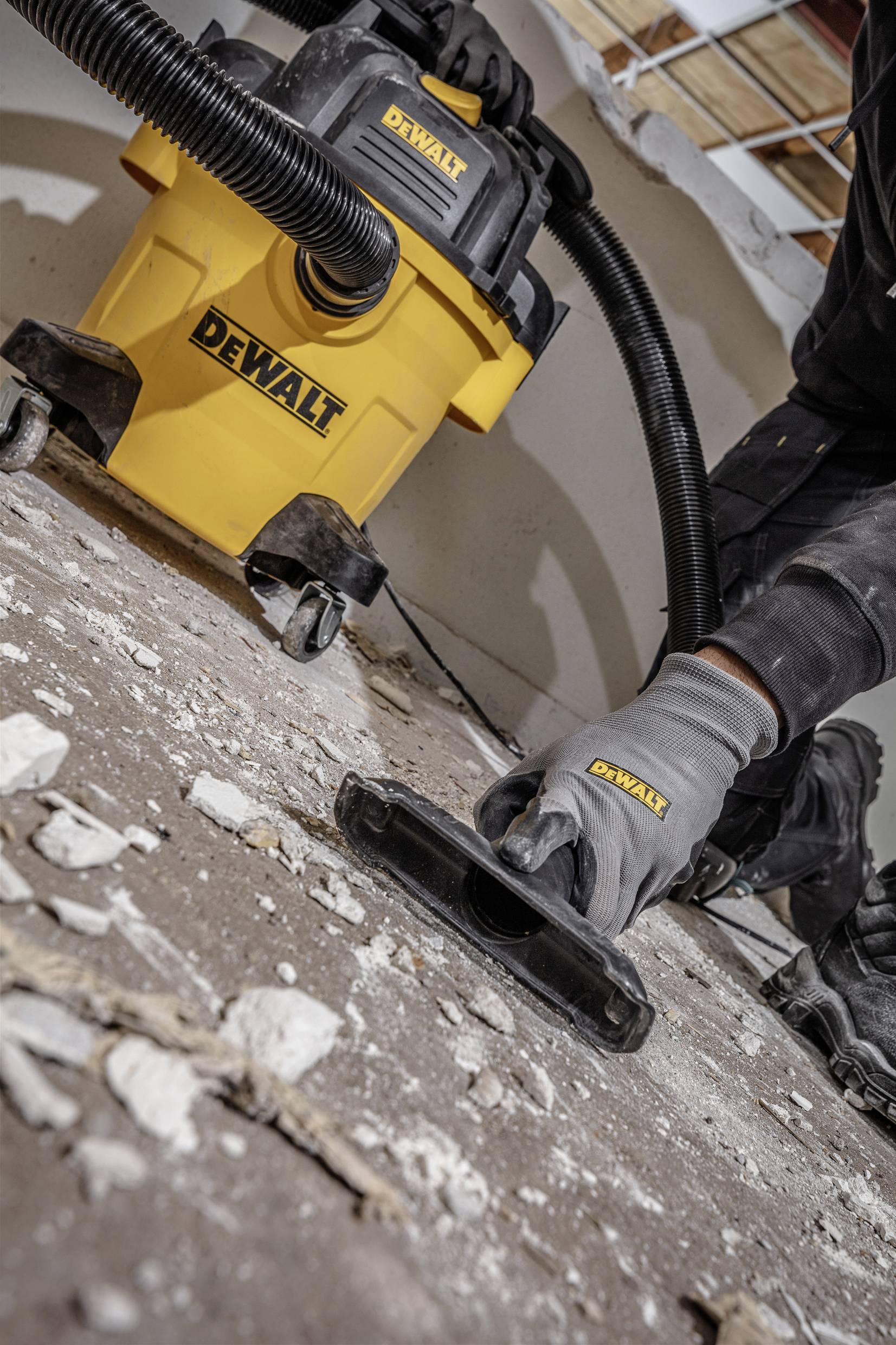 A yellow industrial vacuum cleaner is being used to vacuum building debris from a floor. One person is holding the suction hose.