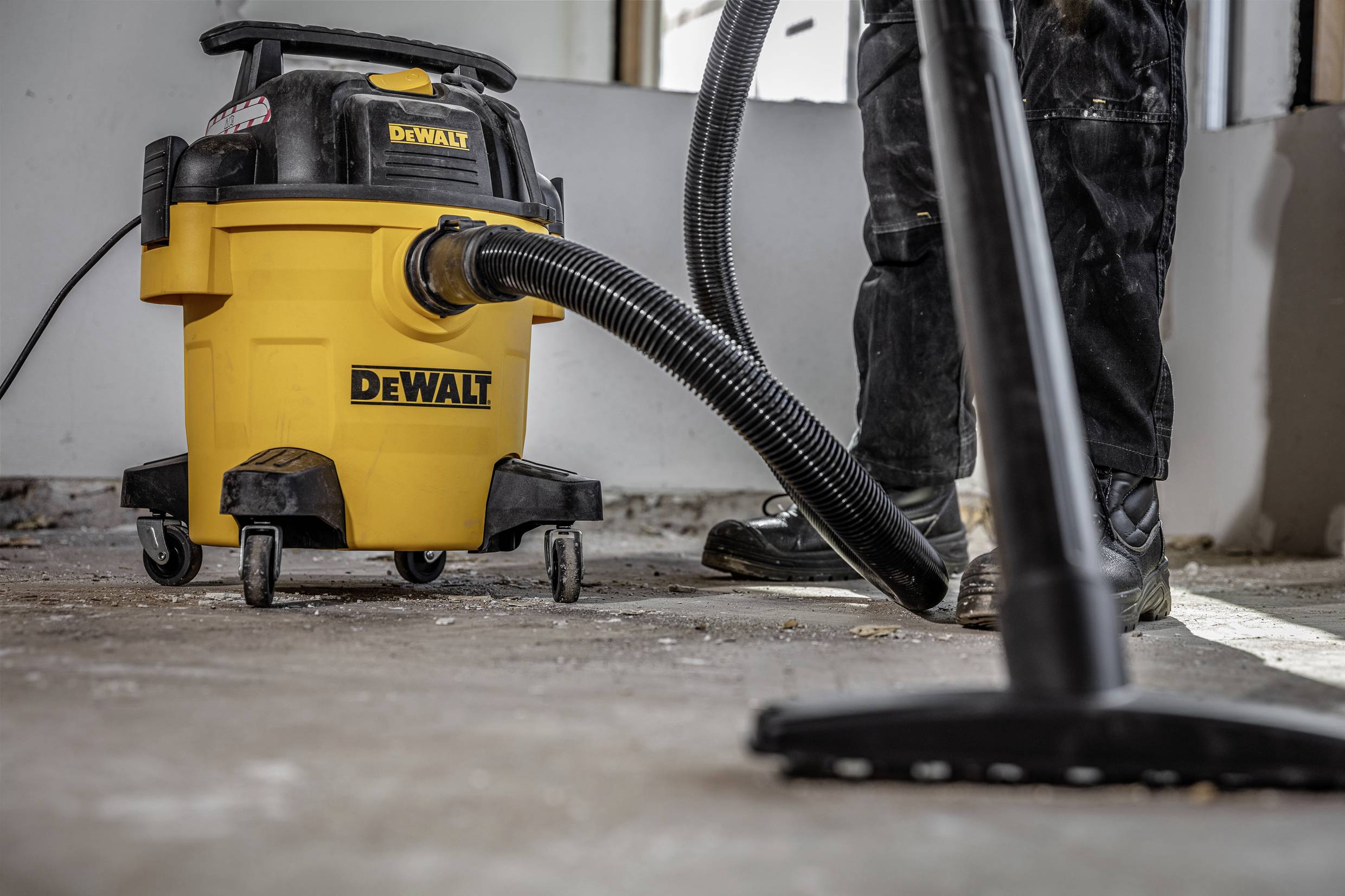 A person is using a yellow industrial vacuum cleaner on a construction site floor. Dust and dirt are being removed.