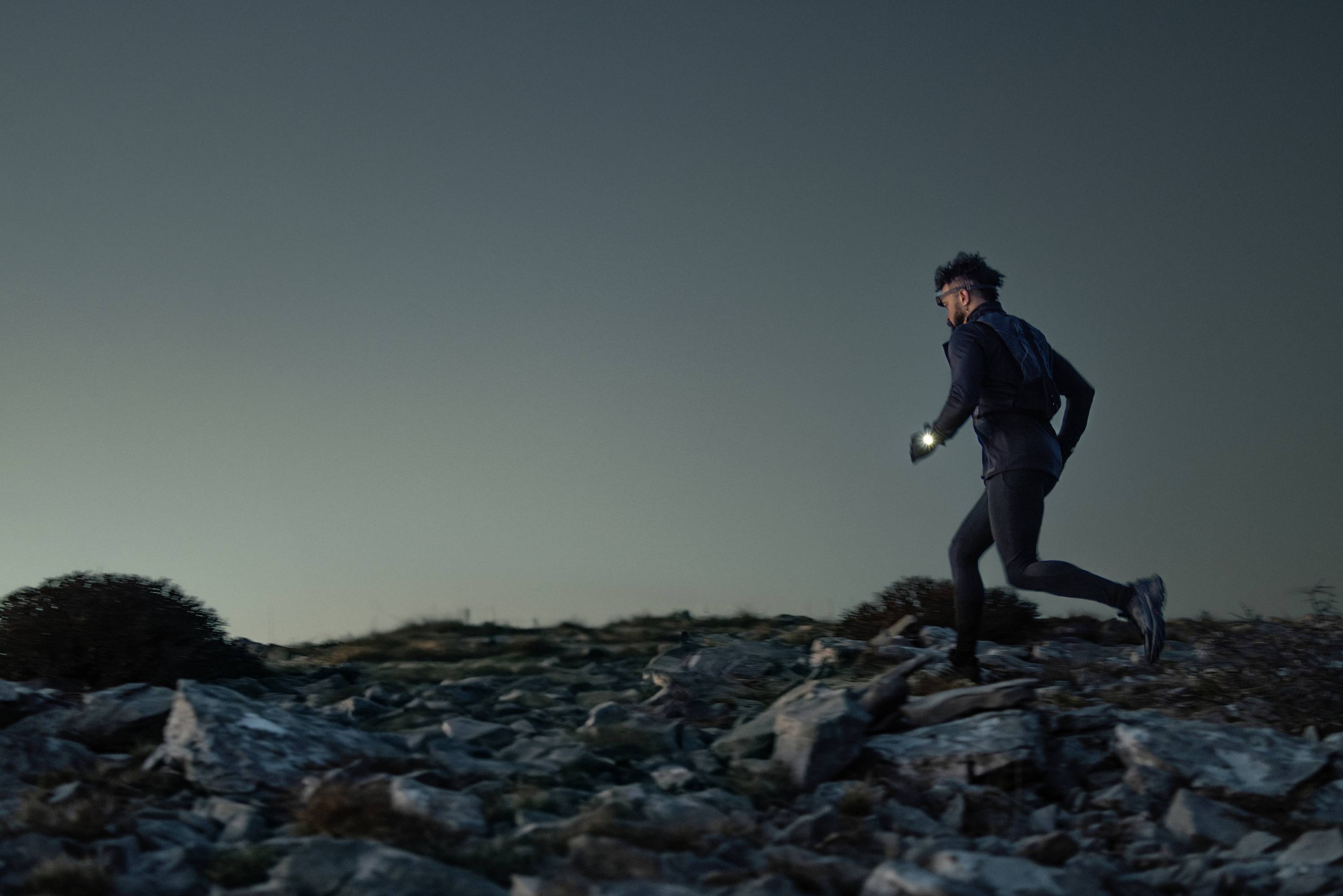 A person is walking across rocky terrain at dusk, wearing a head torch that illuminates the path.