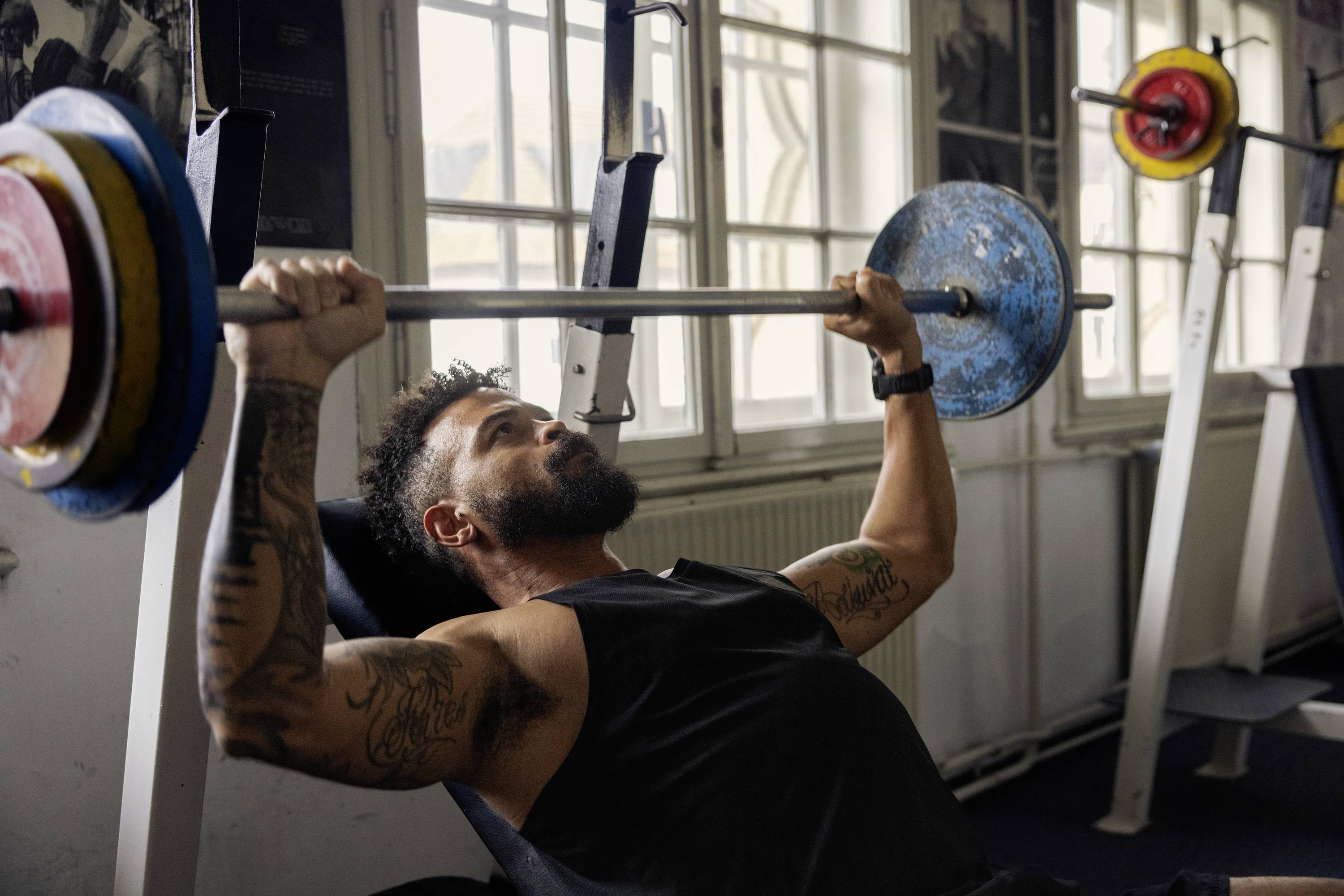 Man with a beard lifts a barbell on a bench in a gym. Tattoos on his arms, focused expression. Equipment room in the background.