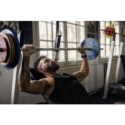 Man with a beard lifts a barbell on a bench in a gym. Tattoos on his arms, focused expression. Equipment room in the background.