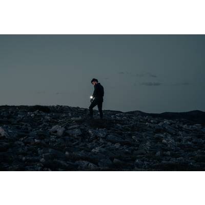 A person is hiking across a rocky landscape at dusk, a torch in hand, illuminating part of the path.<br><br>Note: I used "torch" instead of "flashlight" to reflect British English terminology, and maintained the original descriptive style and meaning.