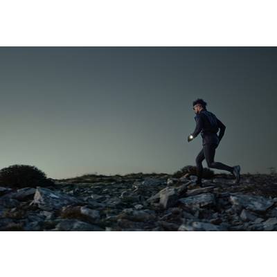 A person is jogging at dusk across rocky terrain, wearing sportswear and holding a torch to illuminate the path.