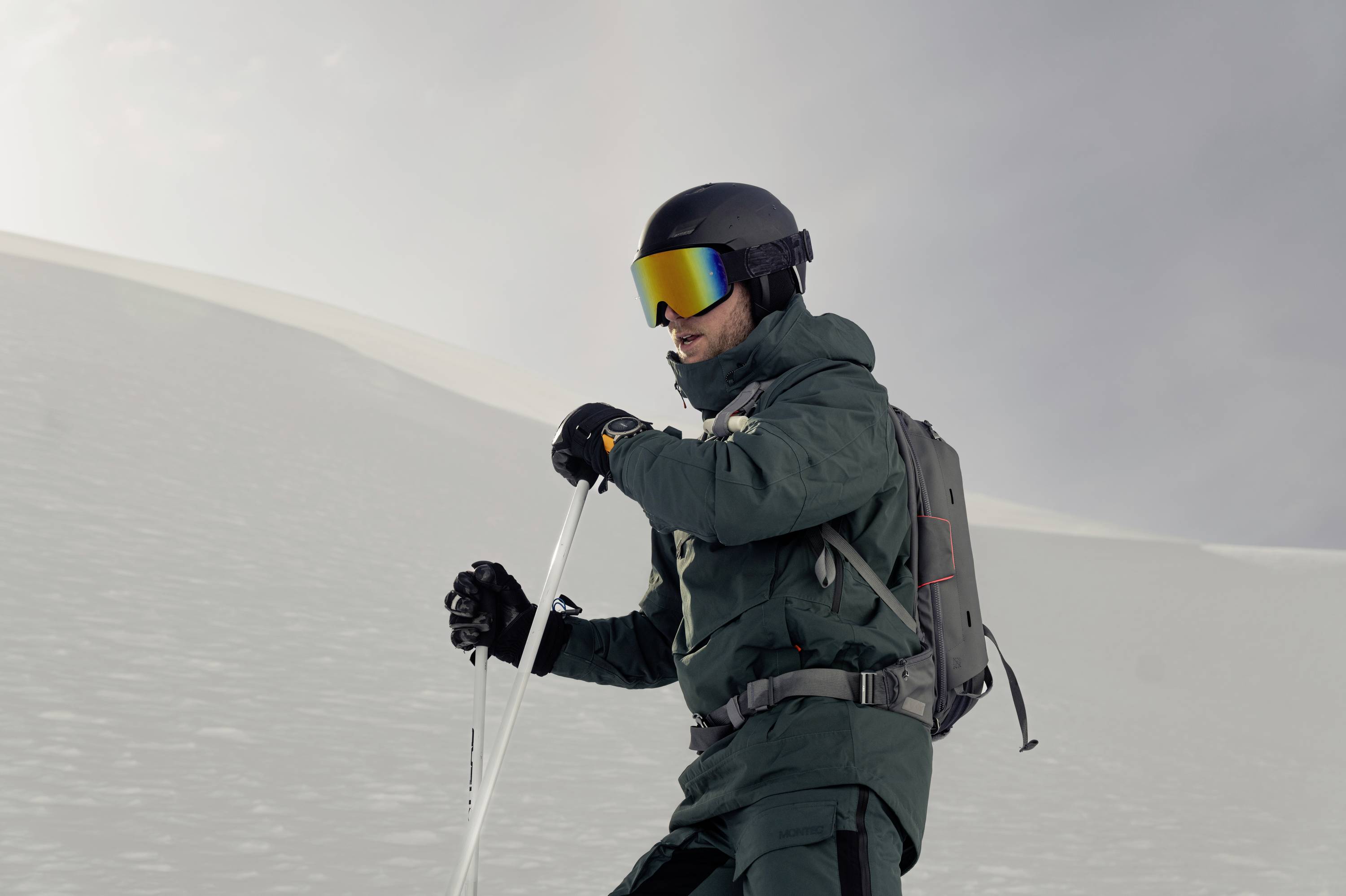 A person in ski gear is skiing down a snow-covered slope, wearing a dark green outfit, helmet, and reflective ski goggles.